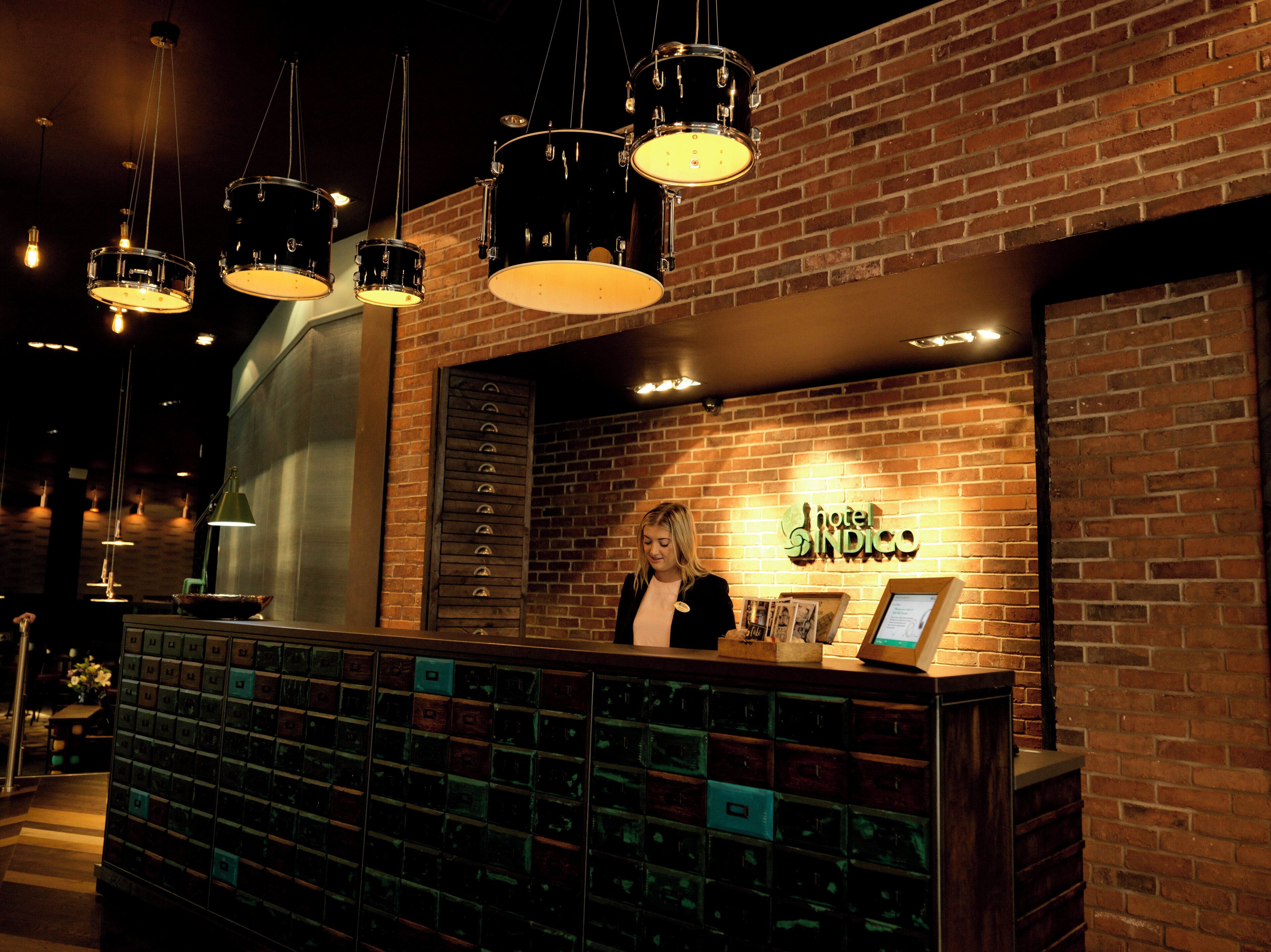 A reception desk with a brick wall background, a sign reading 'Hotel Indigo,' and a series of drawers below the desk.