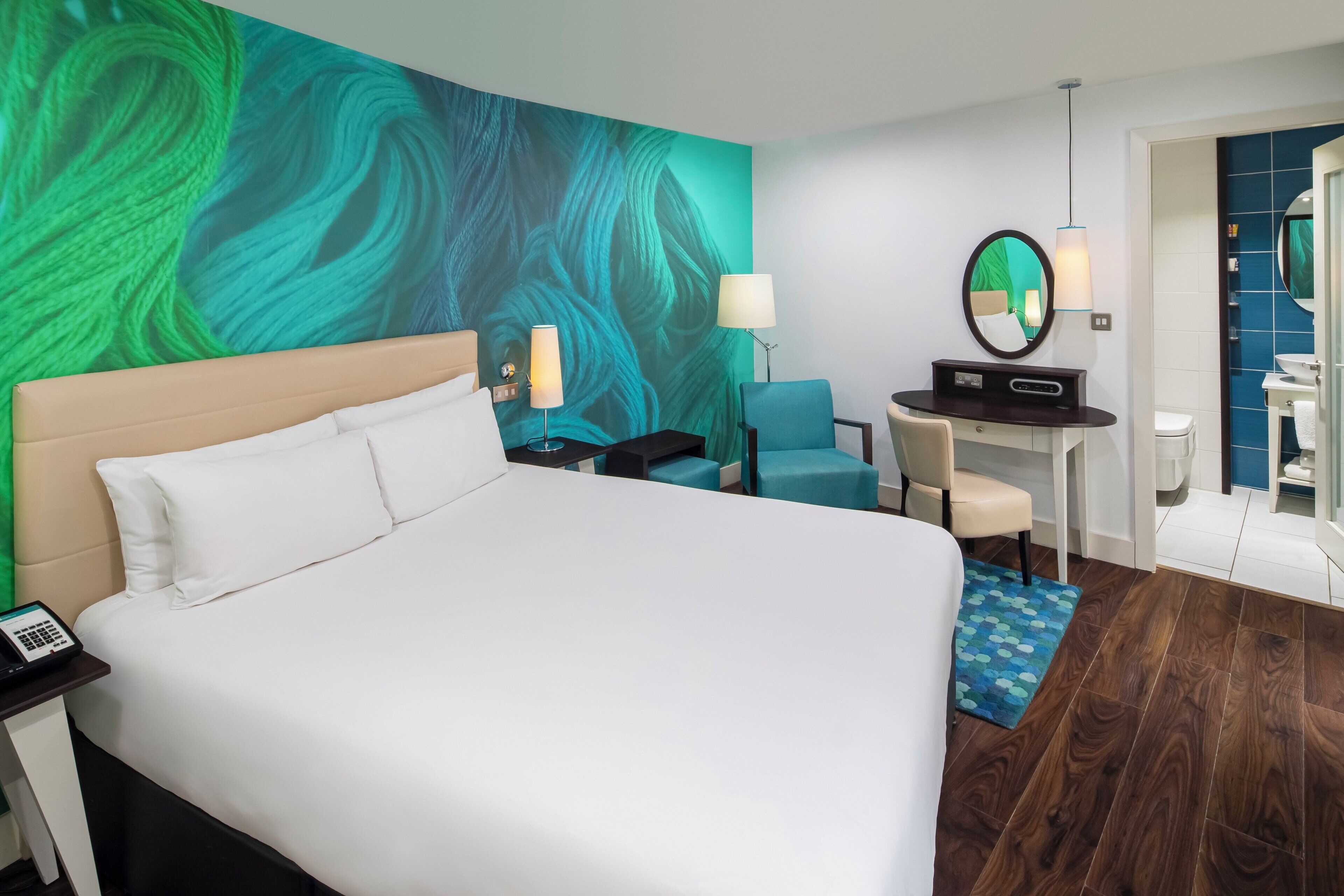 A hotel room with a large bed, a desk with a chair, a bathroom with a mirror, and a wall with a green abstract design.