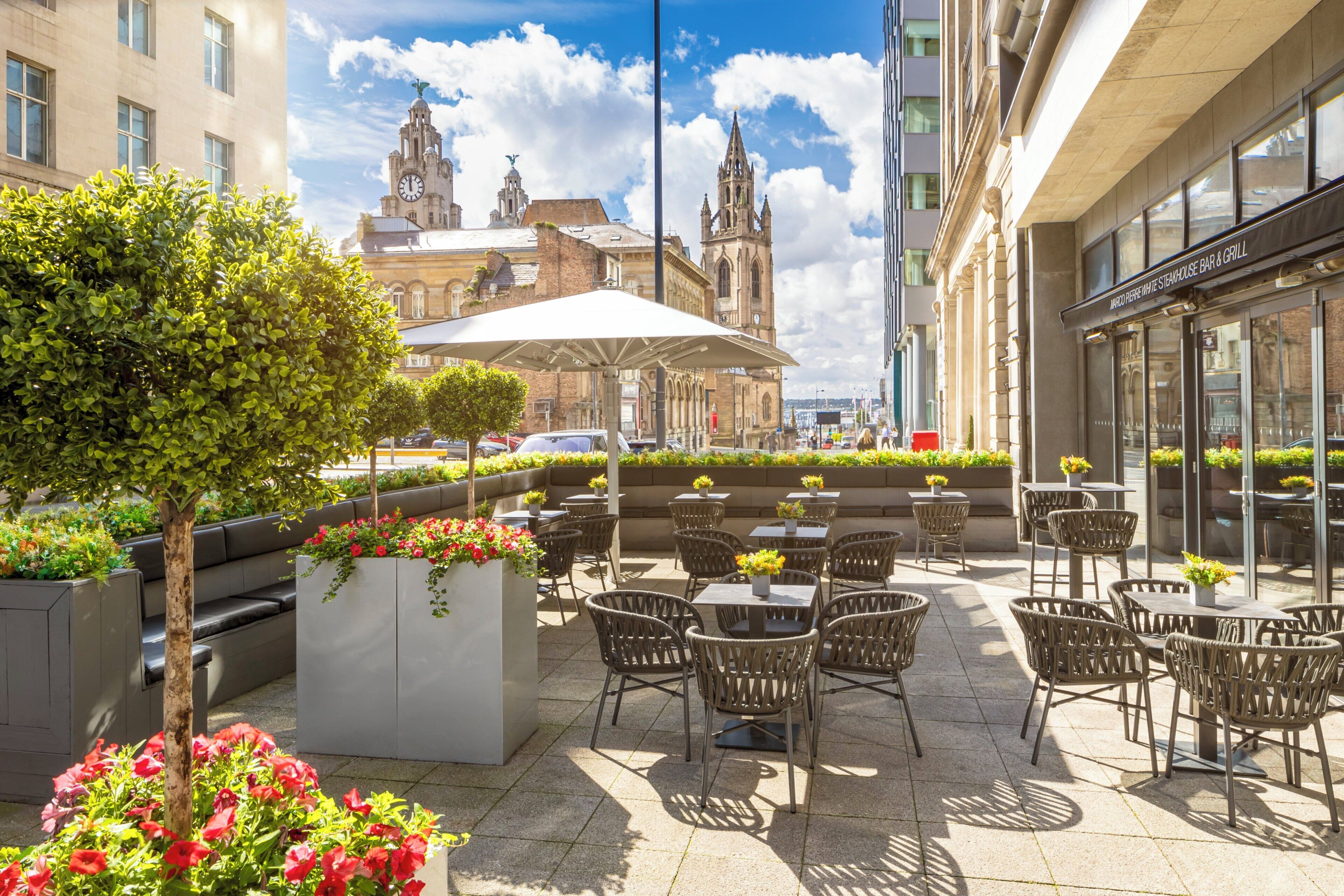 An outdoor patio with wicker chairs and tables, surrounded by flower planters and a large umbrella.