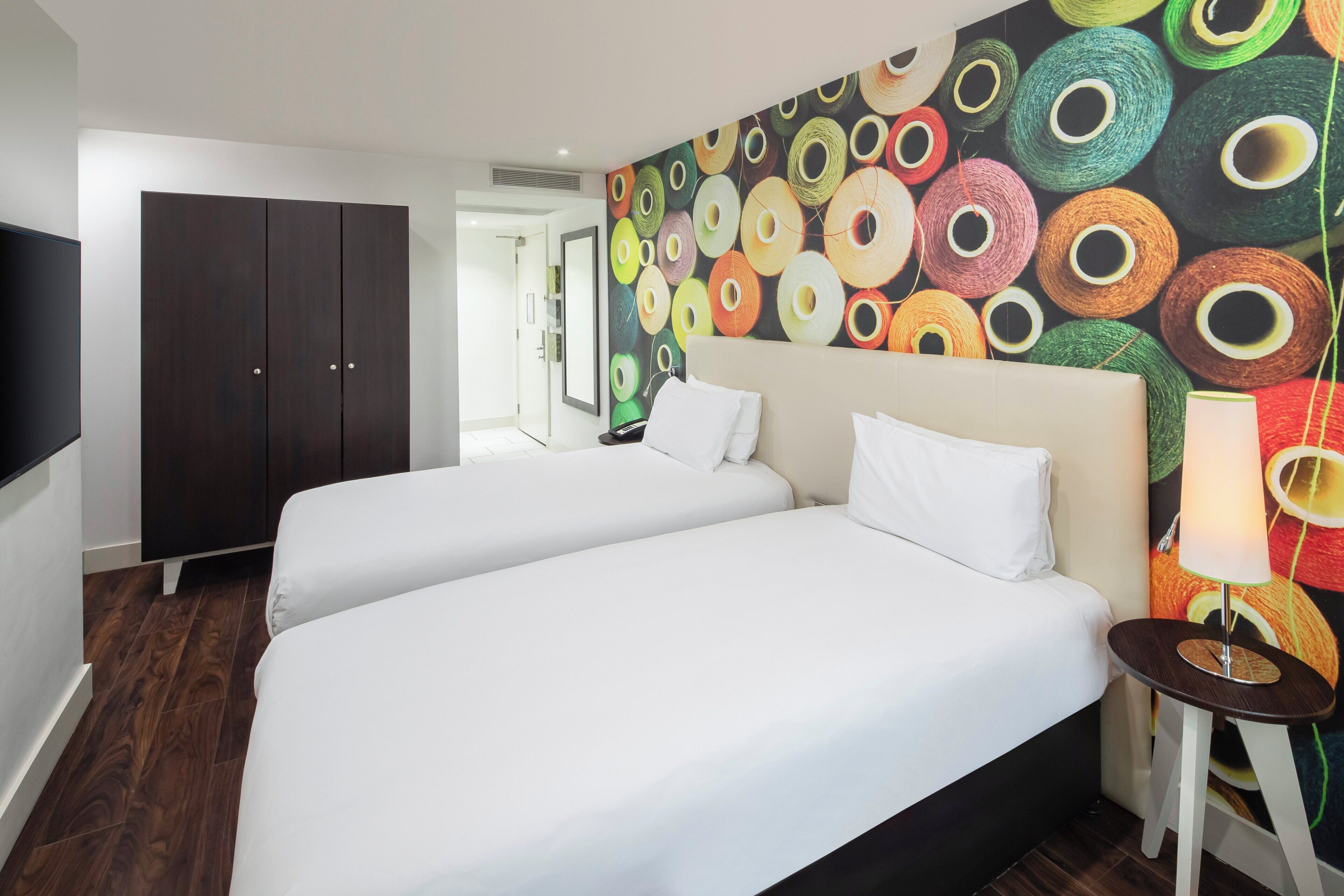 Habitación de hotel con dos camas, un armario de madera oscura, un mural de pared colorido y una mesita de noche pequeña con una lámpara.