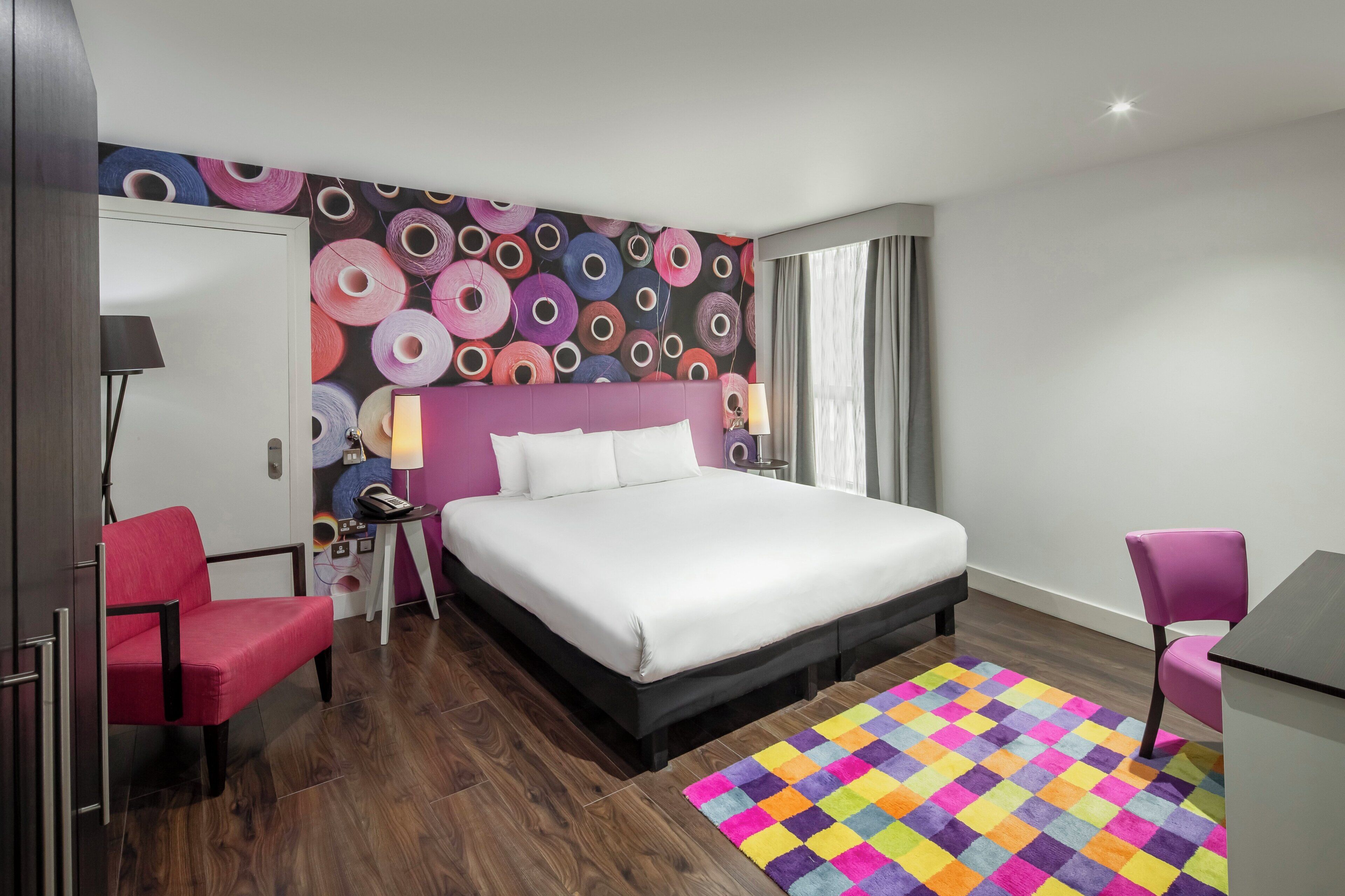 Une chambre d’hôtel avec un grand lit, un bureau, une chaise et un tapis aux couleurs vives.