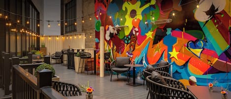 An outdoor seating area with tables and chairs, a mural on the wall, and string lights.
