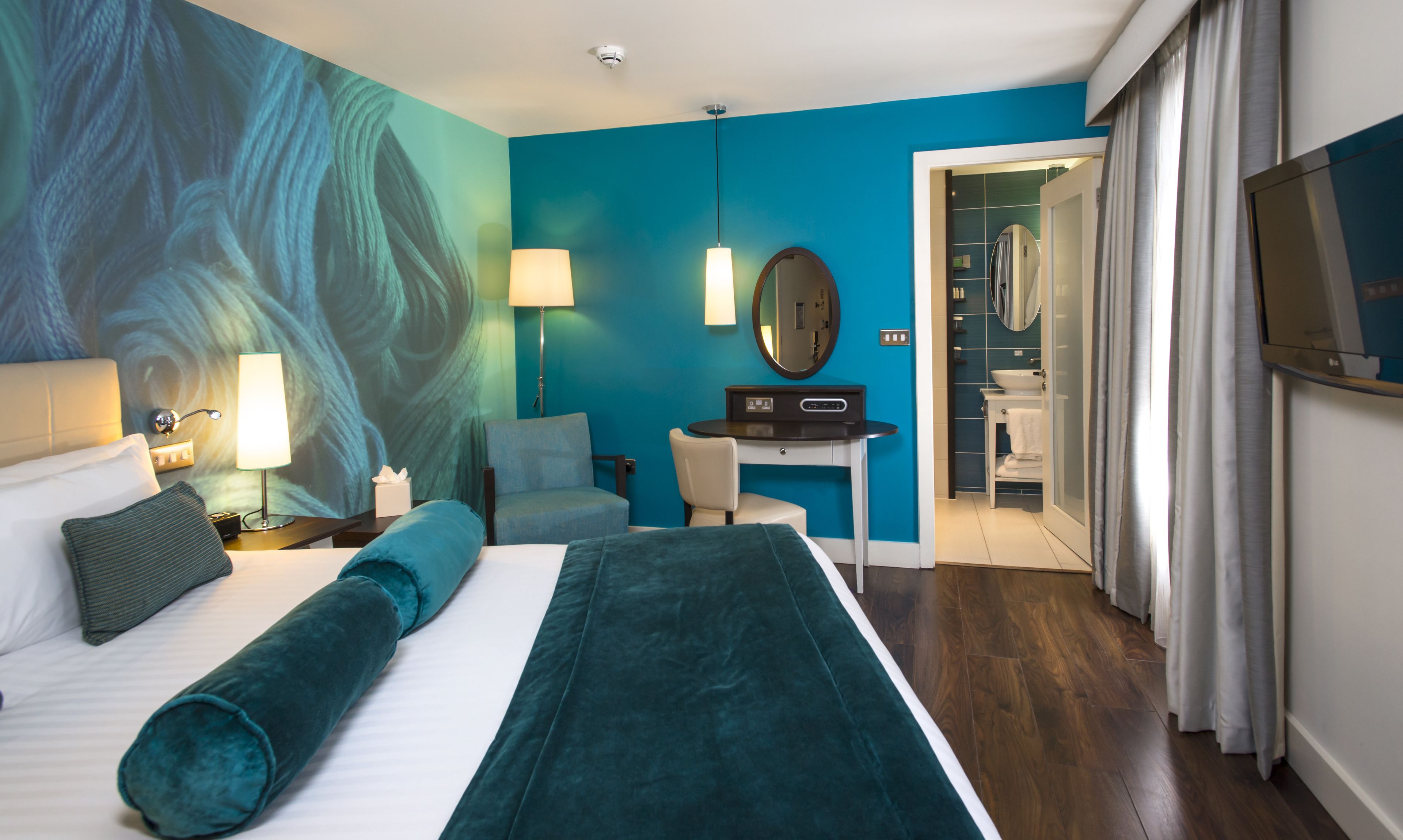 A hotel room with a large bed, a desk, a chair, a mirror, and a bathroom visible through an open door.