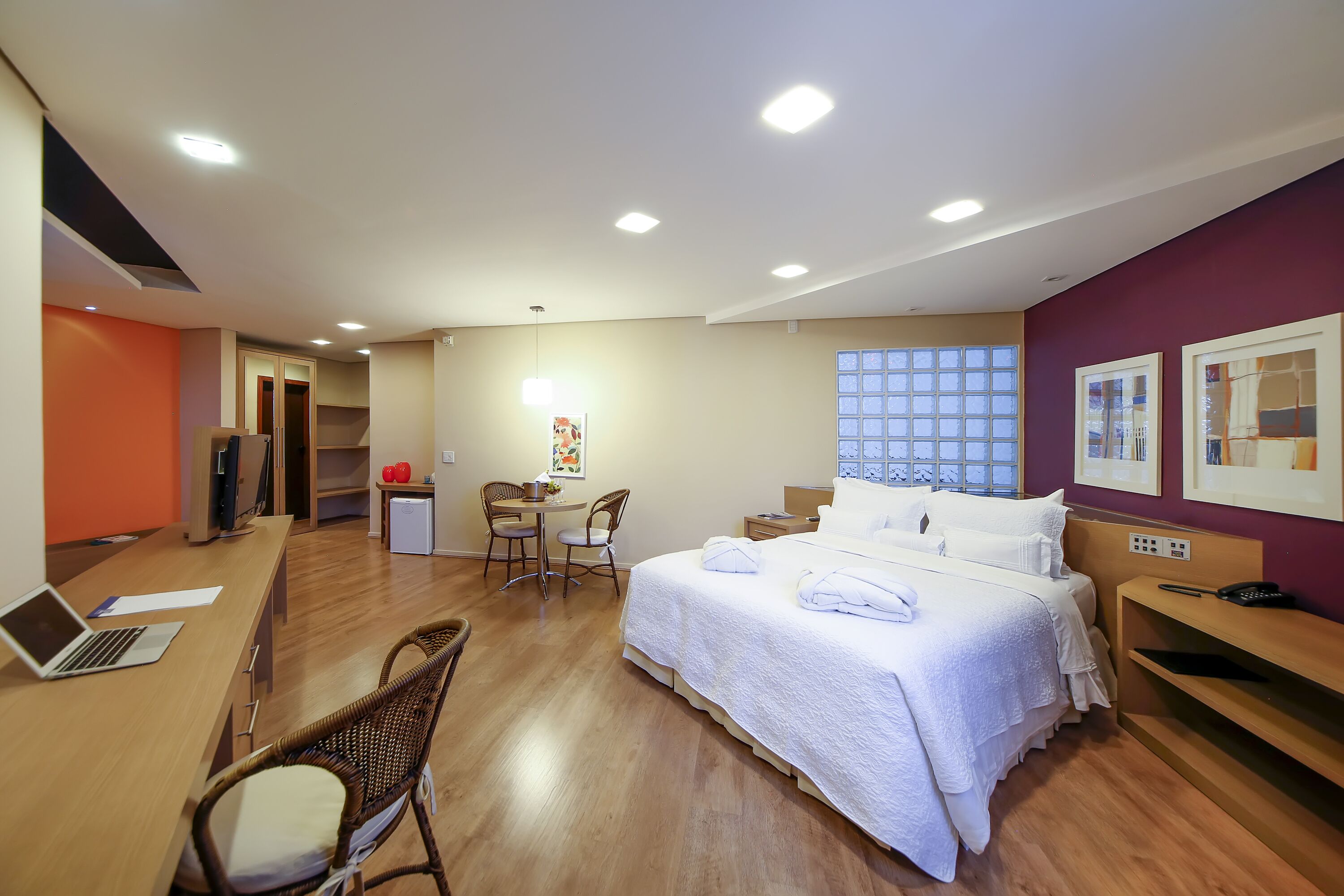 A hotel room with a bed, a desk with a laptop, a chair, a small table with a lamp, and a window with blinds.