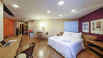 A hotel room with a bed, a desk with a laptop, a chair, a small table with a lamp, and a window with blinds.