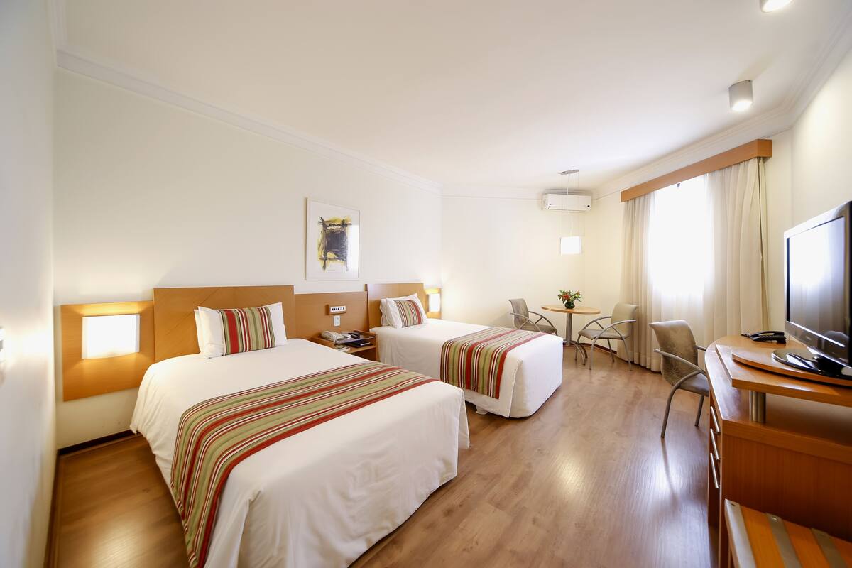 Une chambre d’hôtel avec deux lits, un bureau avec une télévision, une chaise, une petite table avec une plante et une fenêtre avec des rideaux.