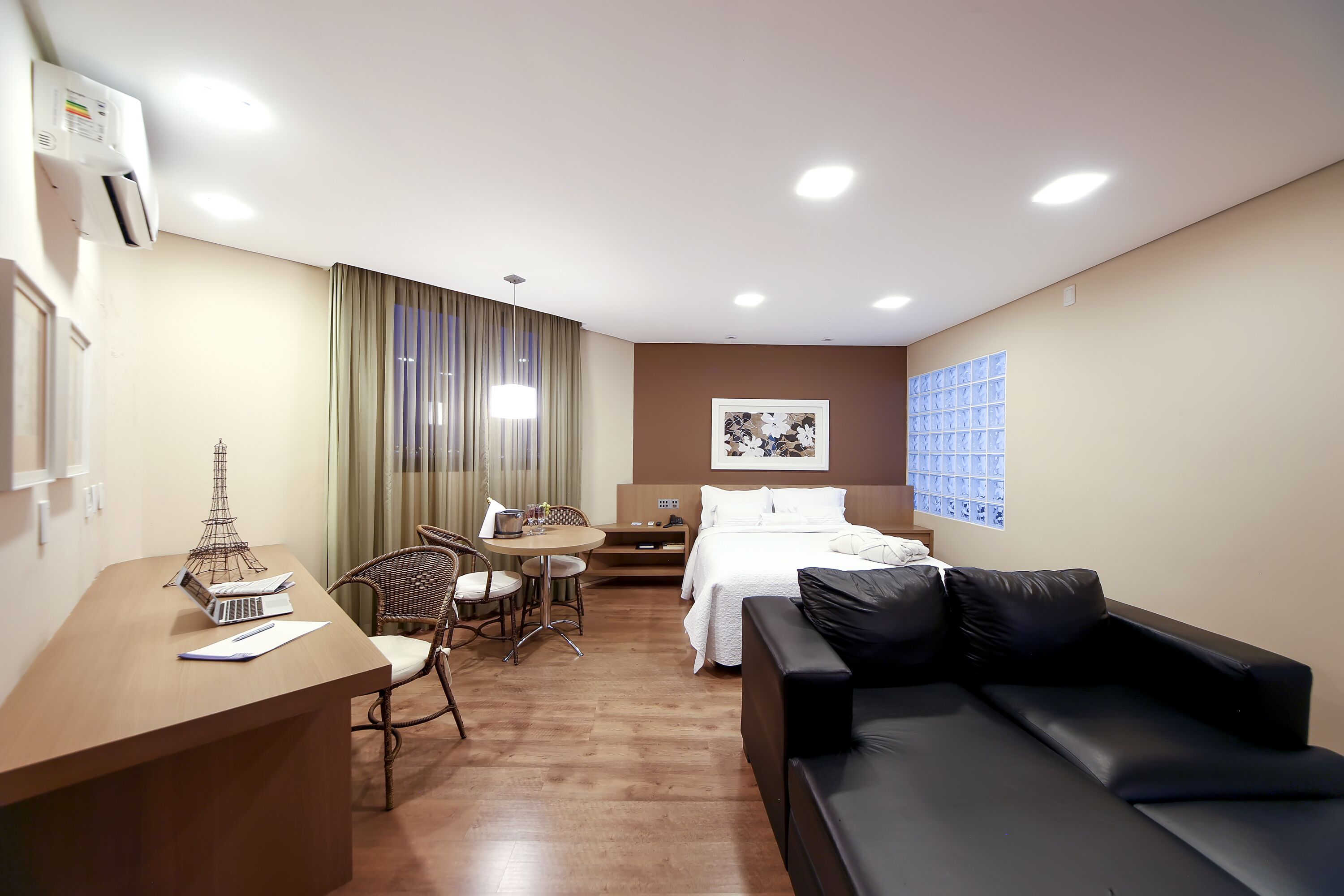 Habitación de hotel con cama, sofá de cuero negro, escritorio de madera con una maqueta de la Torre Eiffel, mesa de comedor con sillas y ventana con persianas.