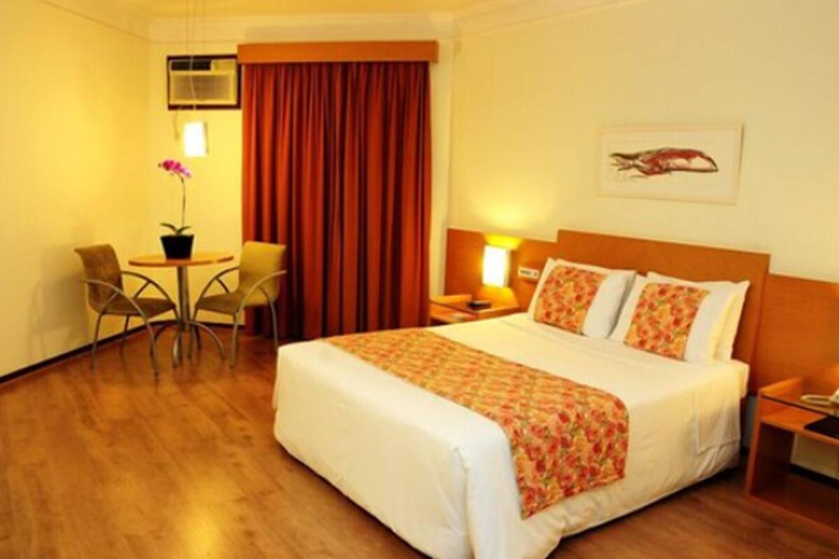 Une chambre d’hôtel comprenant un lit, un bureau avec une chaise, une petite table avec un pot de plante et un climatiseur fixé au mur.