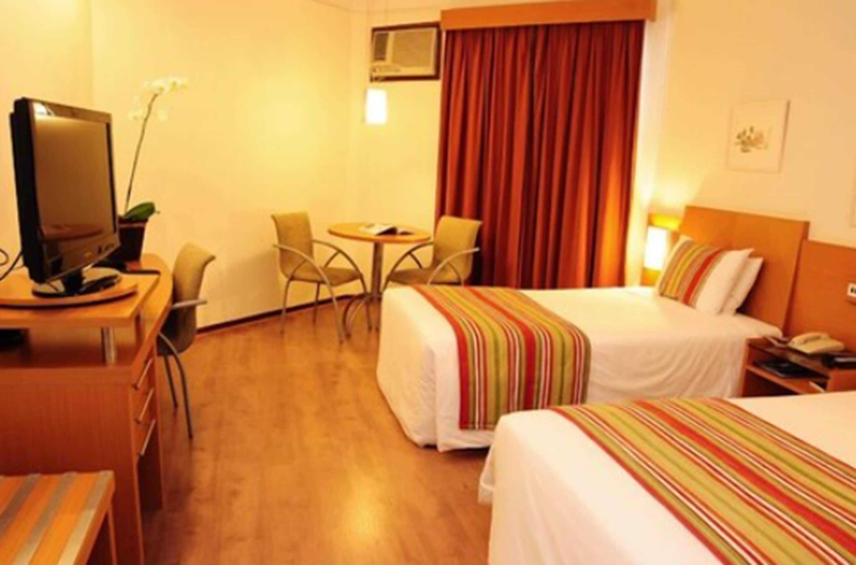 Quarto de hotel com duas camas, uma secretária com computador, uma cadeira e uma televisão.