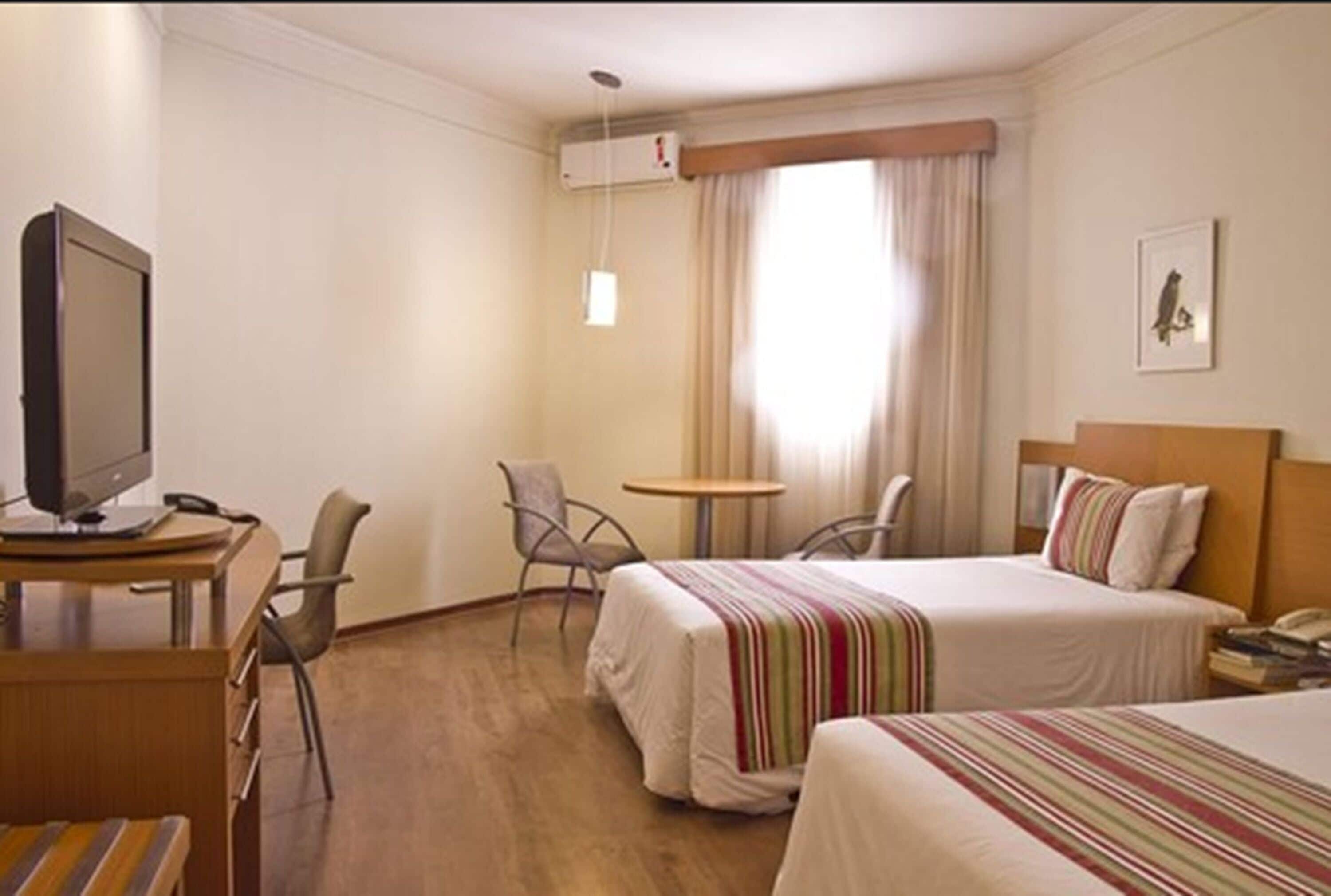 Habitación de hotel con dos camas, un escritorio con computadora, una silla, una mesita y una ventana con cortinas.