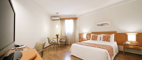 A hotel room with a bed, a desk with a computer, a chair, a small table with flowers, and a window with curtains.