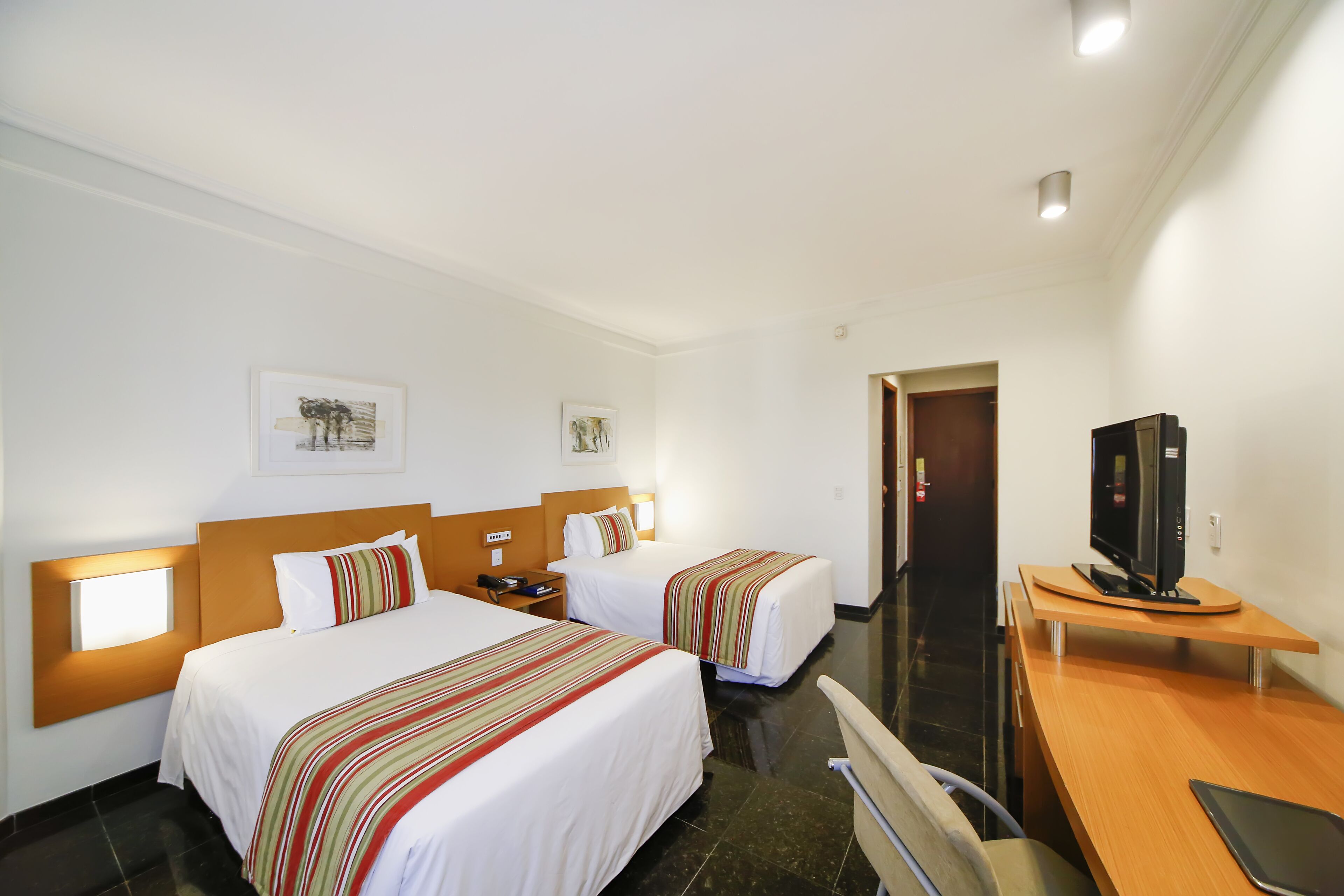 A hotel room with two beds, a desk, a television, and artwork on the walls.