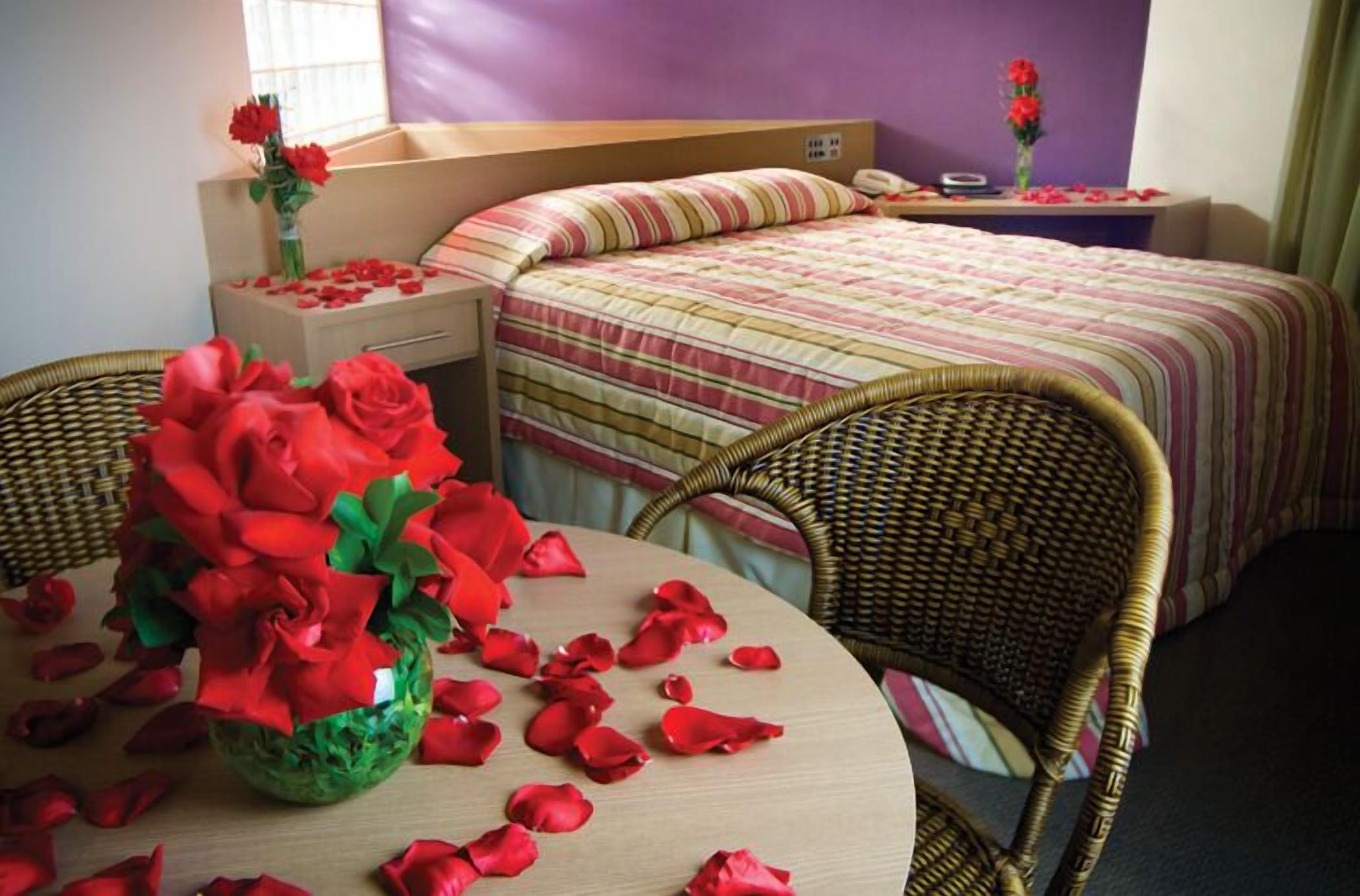 A hotel room with two beds, a wicker chair, a table with a vase of red roses, and a window with a view.
