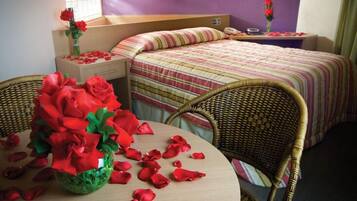 A hotel room with two beds, a wicker chair, a table with a vase of red roses, and a window with a view.