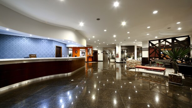A modern hotel lobby with a curved reception desk, a seating area with a bench and a coffee table, and a decorative plant. - Grand Hotel Royal Sorocaba by Atlantica (Sorocaba)