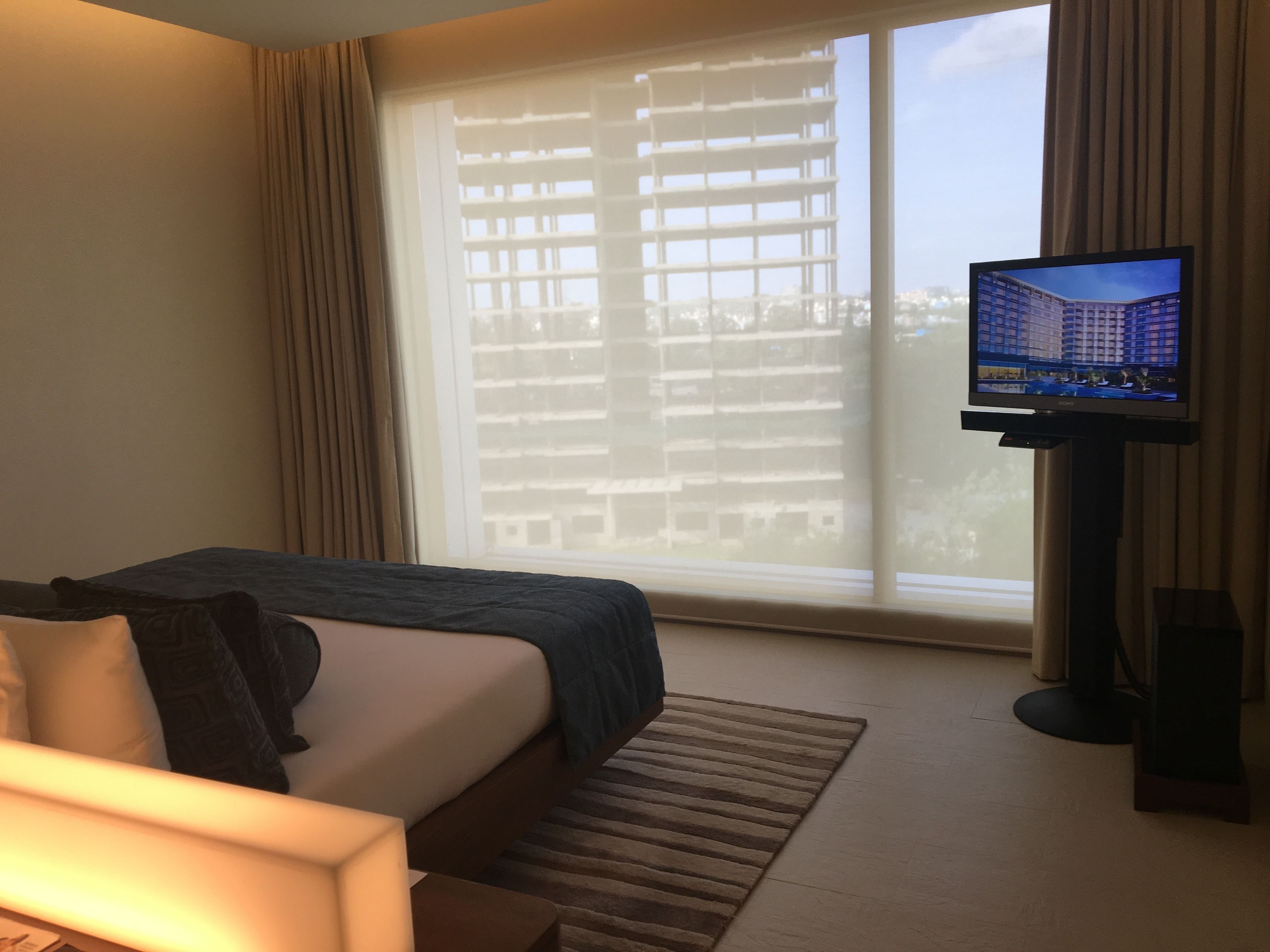 Habitación de hotel con cama, televisión y un amplio ventanal con vistas a edificios.