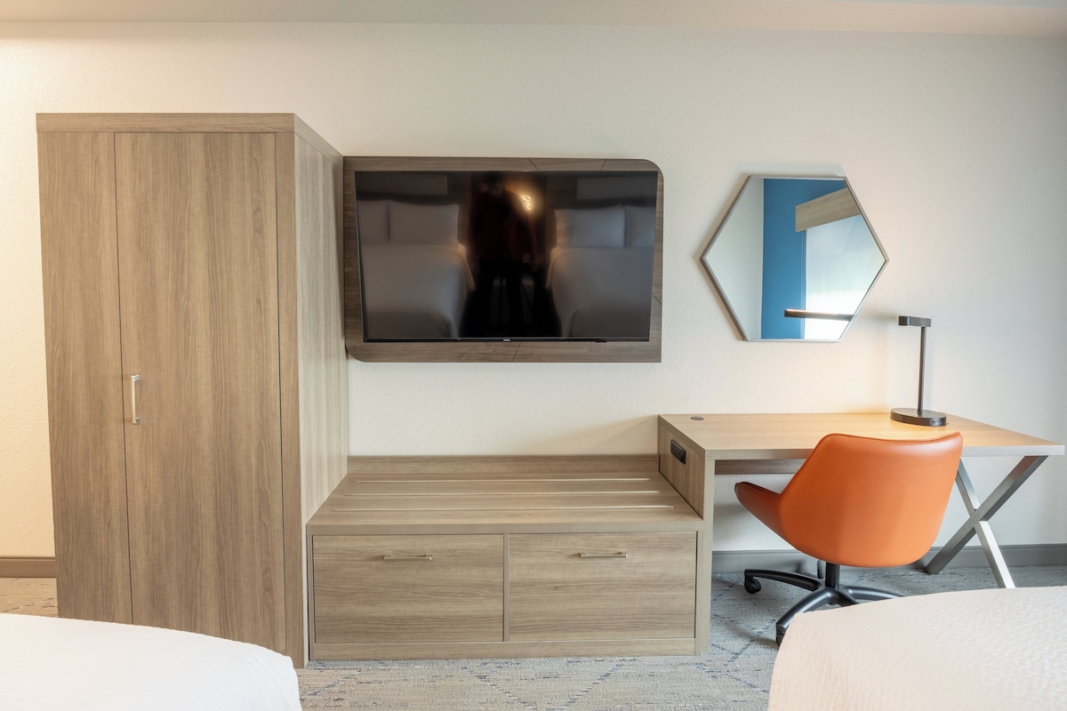 Quarto de hotel com cama, secretária, cadeira, televisão, guarda-roupa e espelho.