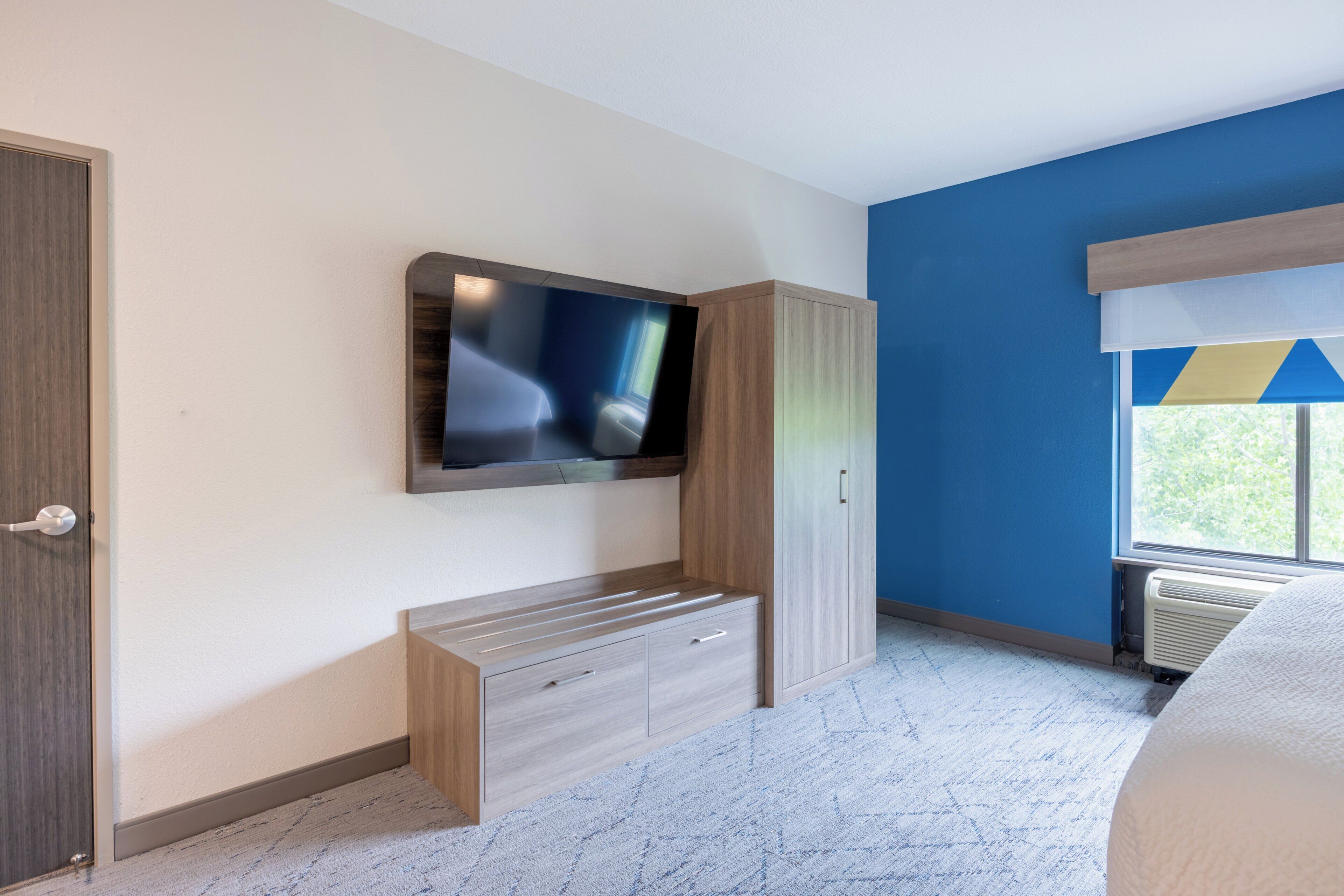A bedroom with a flat-screen TV, a wooden bench, a wardrobe, and a window with blinds.