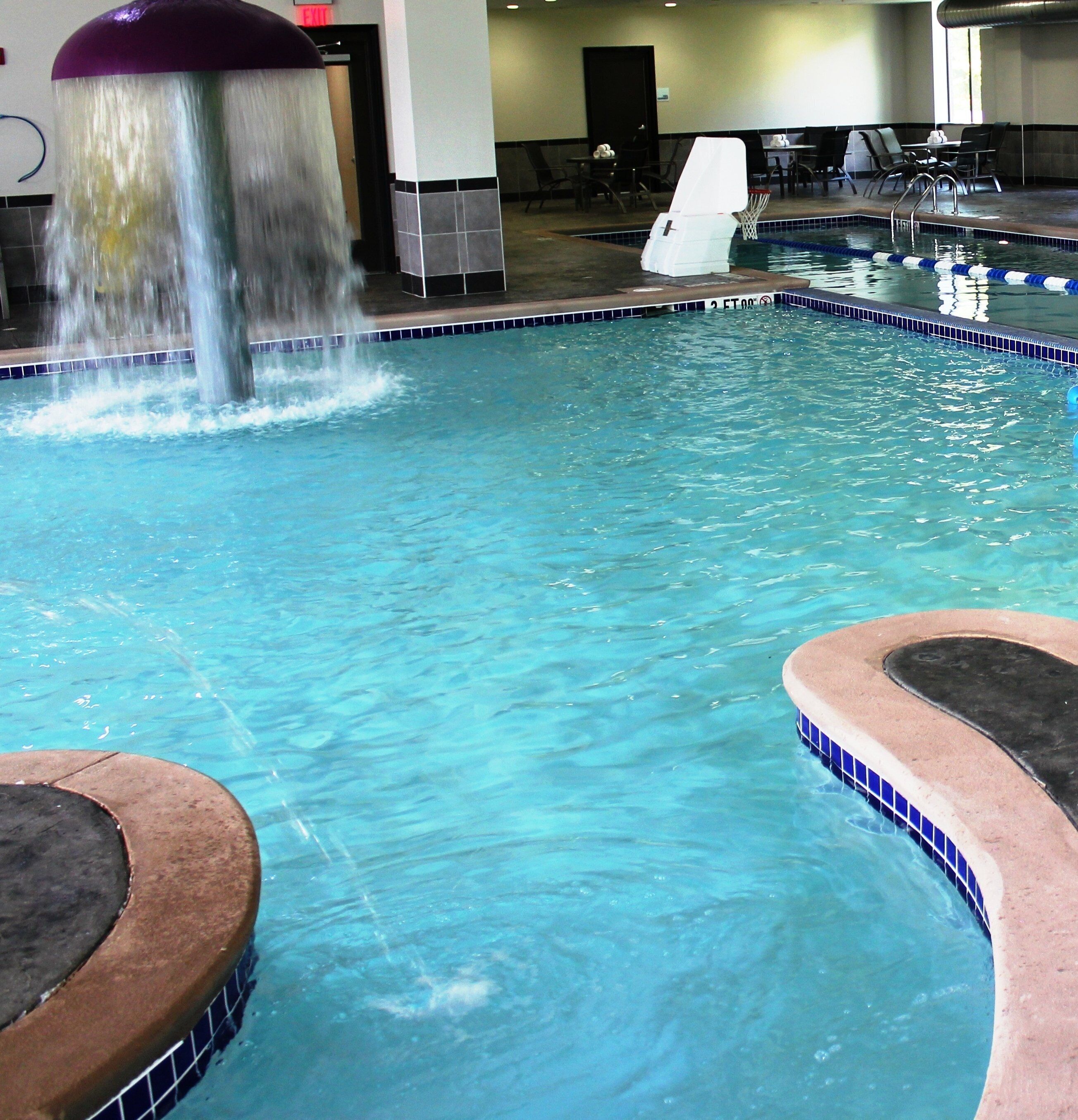 An indoor swimming pool with a water feature, tiled edges, and a seating area with tables and chairs.