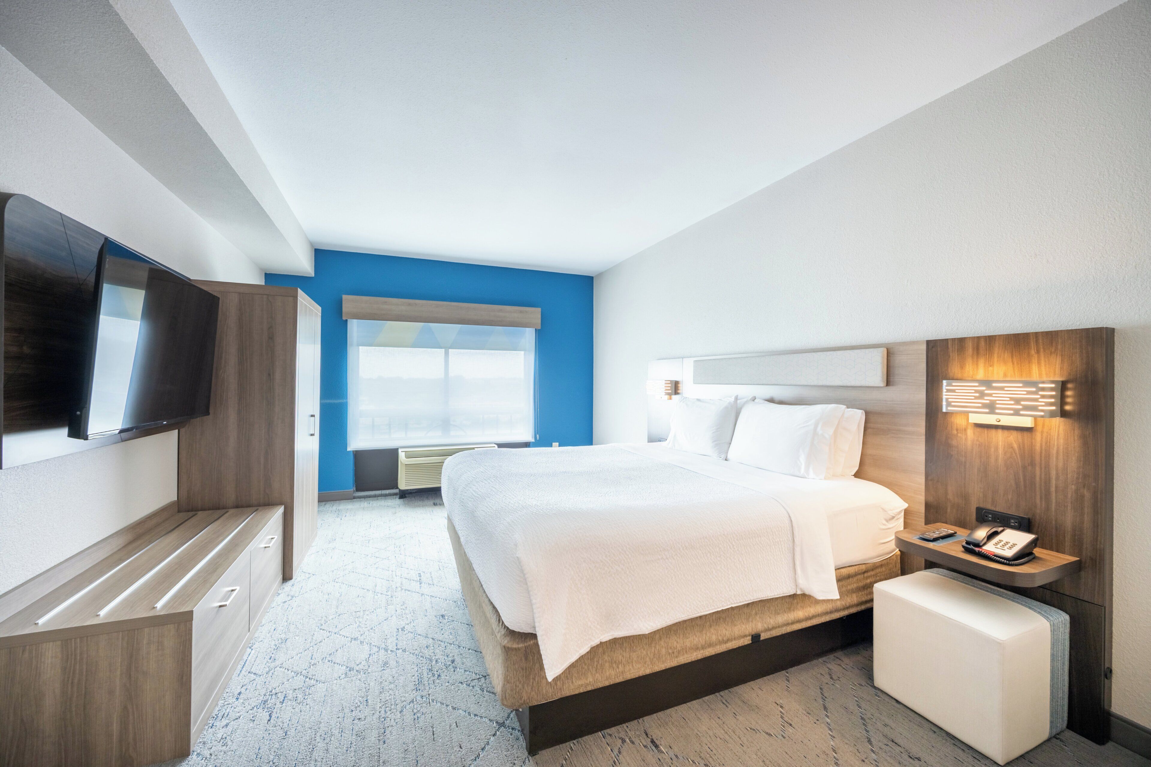 Ein modernes Hotelzimmer mit Bett, Fernseher, Nachttisch mit Telefon und Fenster mit Jalousien.