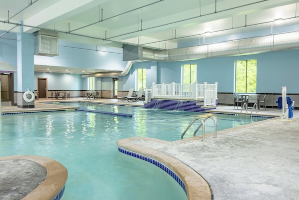 An indoor swimming pool with clear blue water, a diving board, and a seating area with tables and chairs.
