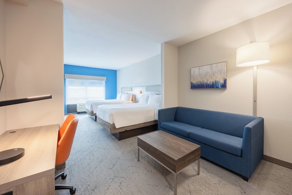 Quarto de hotel com cama, um sofá azul, uma mesa com cadeira, luminária e um quadro na parede.