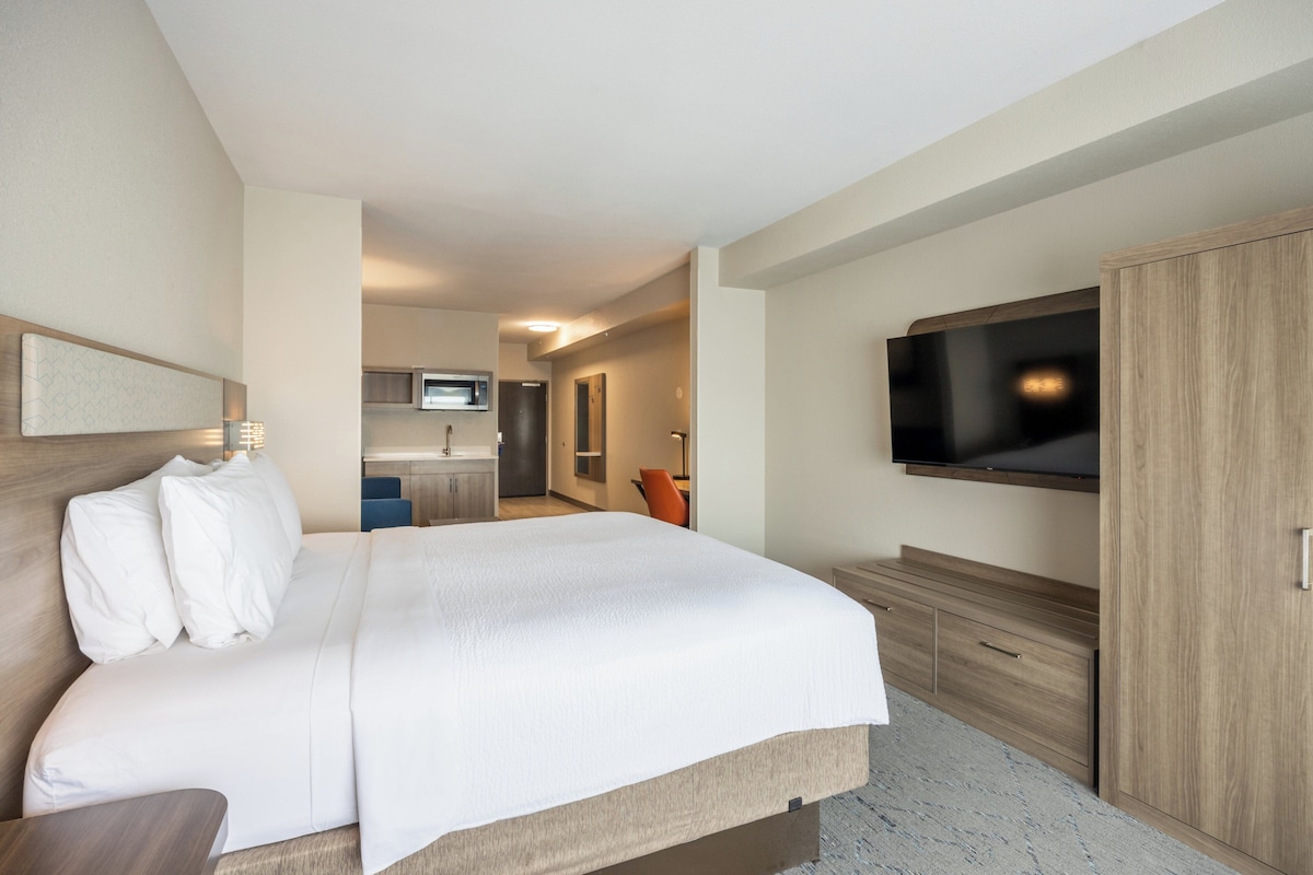 Quarto de hotel com cama, televisão, guarda-roupa e uma kitchenette pequena.