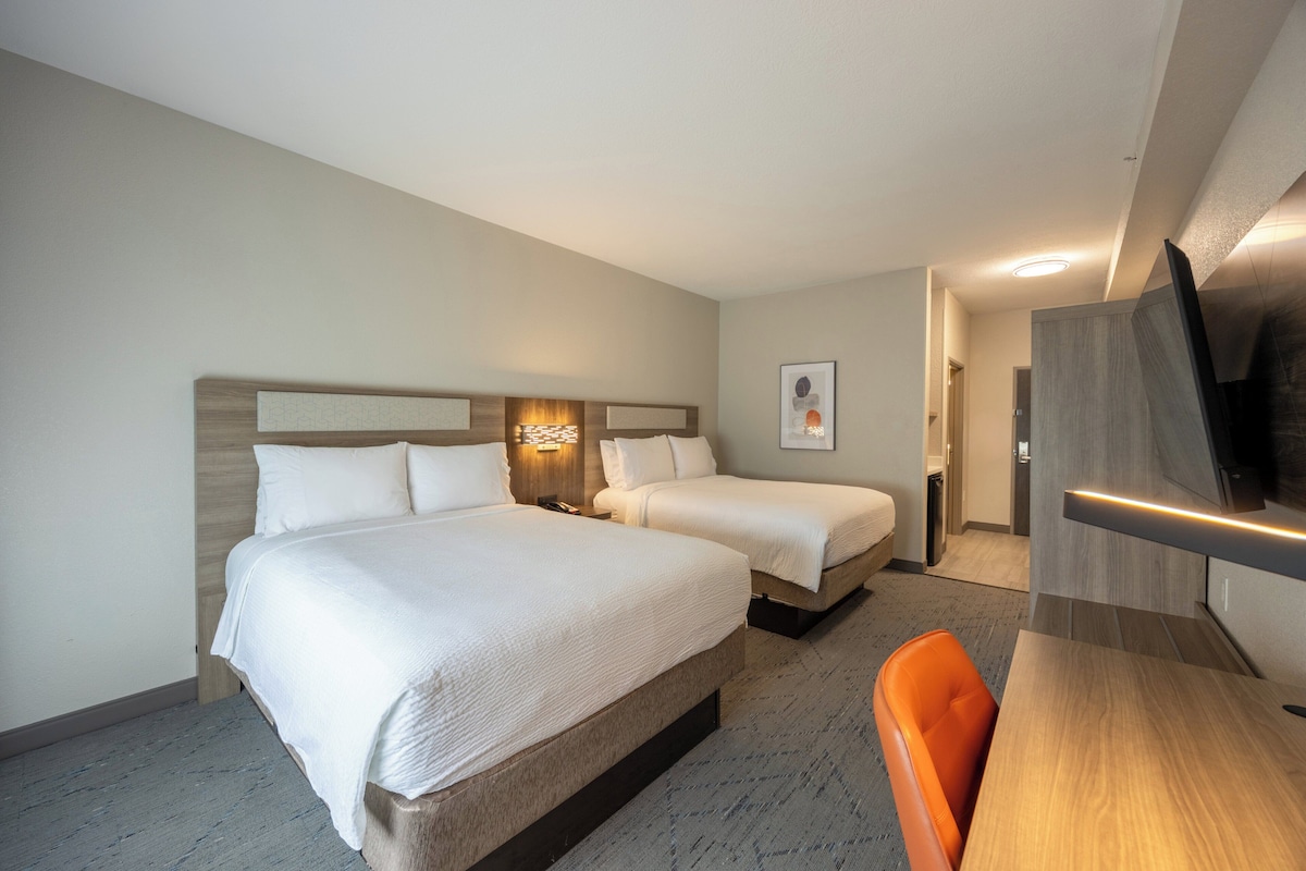 Quarto de hotel com duas camas, uma escrivaninha, uma cadeira, uma televisão e um quadro pendurado na parede.