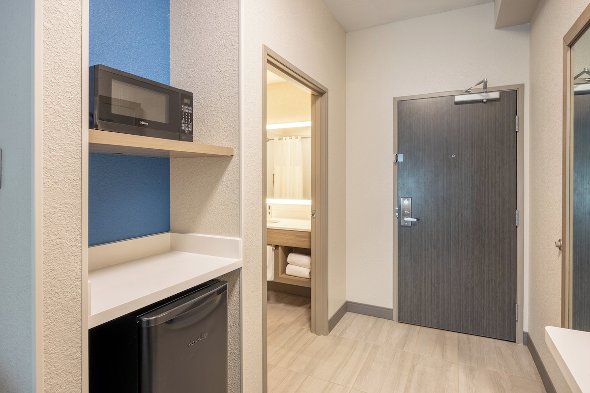 Quarto de hotel pequeno com micro-ondas, mini-geladeira, prateleira de madeira, porta e banheiro visível através de uma porta aberta.