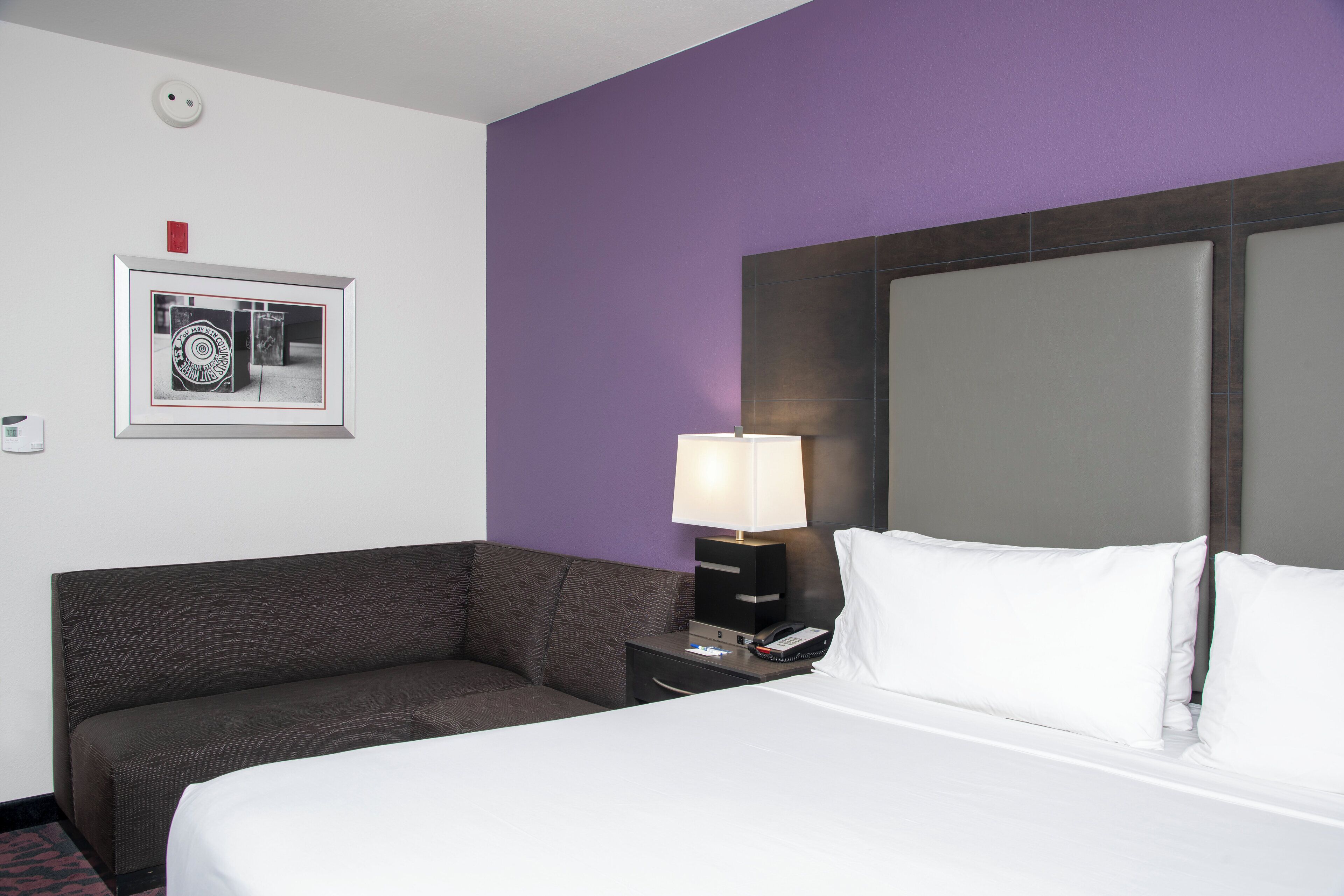 Quarto de hotel com uma cama, mesa de cabeceira, abajur, sofá e um quadro na parede.
