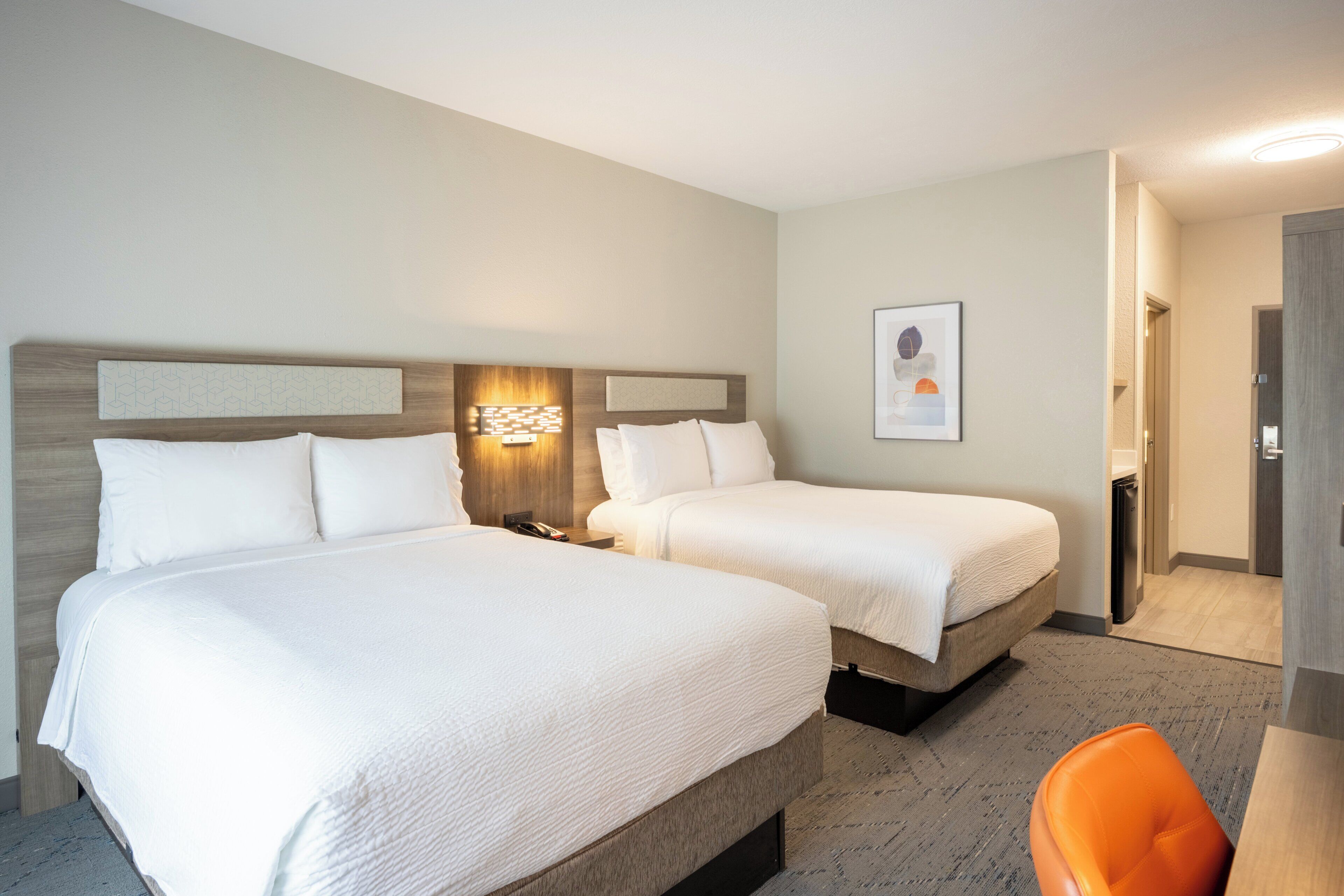 Quarto de hotel com duas camas, uma escrivaninha, uma cadeira, uma luminária na parede e um quadro emoldurado.