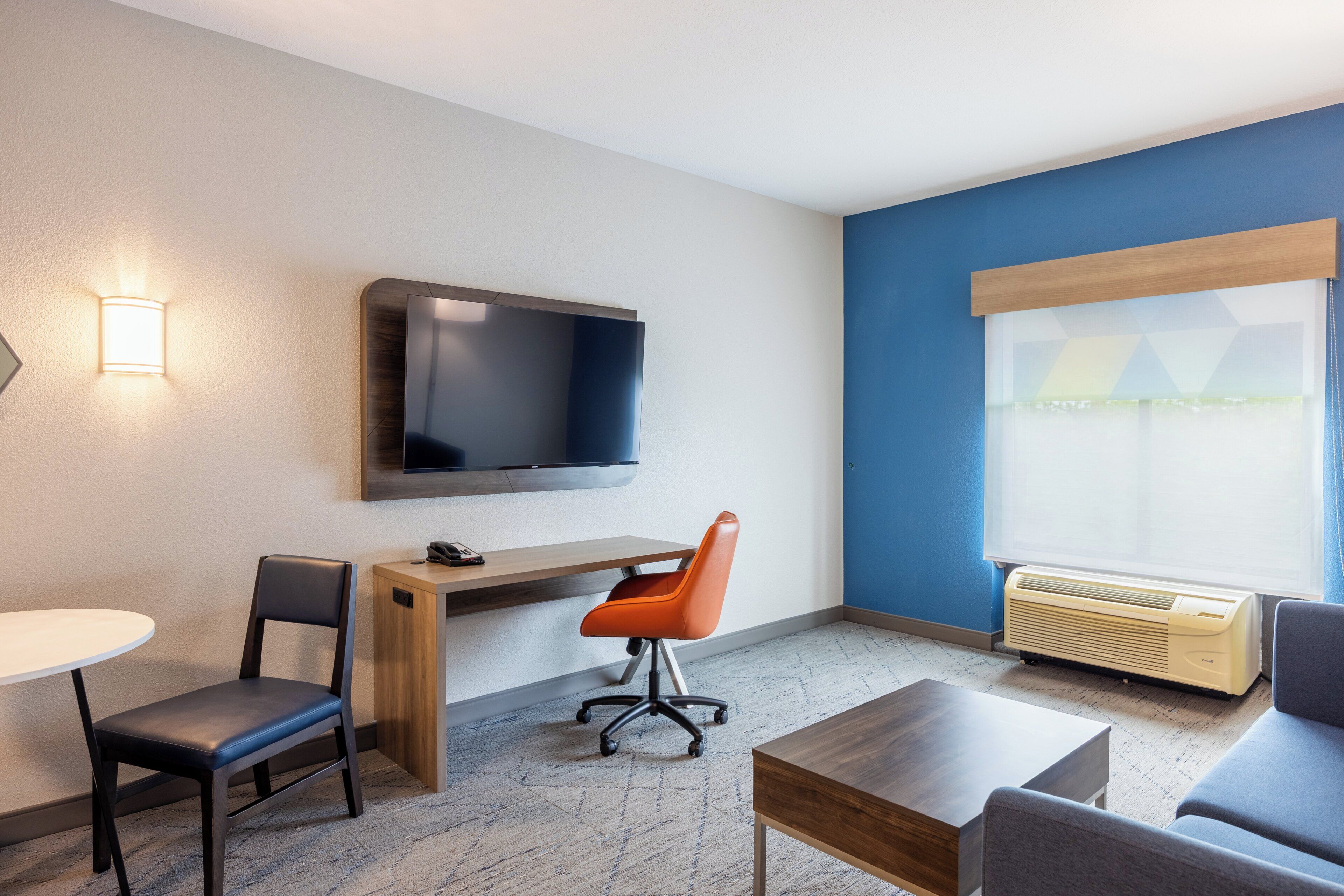 Habitación de hotel con televisión de pantalla plana en la pared, escritorio con silla, sofá, mesita y ventana con aire acondicionado.