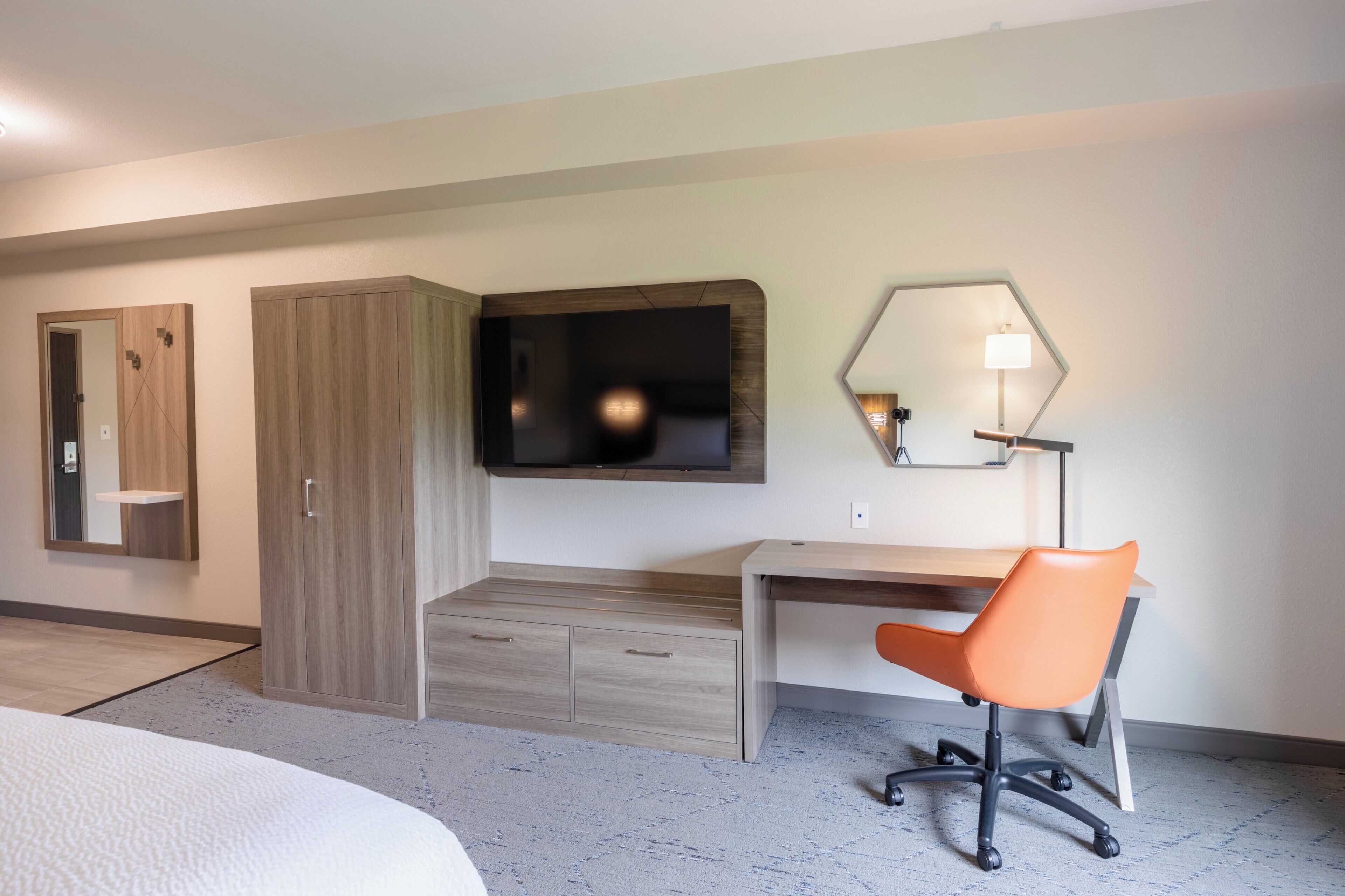 Une chambre d’hôtel avec un lit, un bureau avec une chaise orange, une télévision, un miroir et une armoire.
