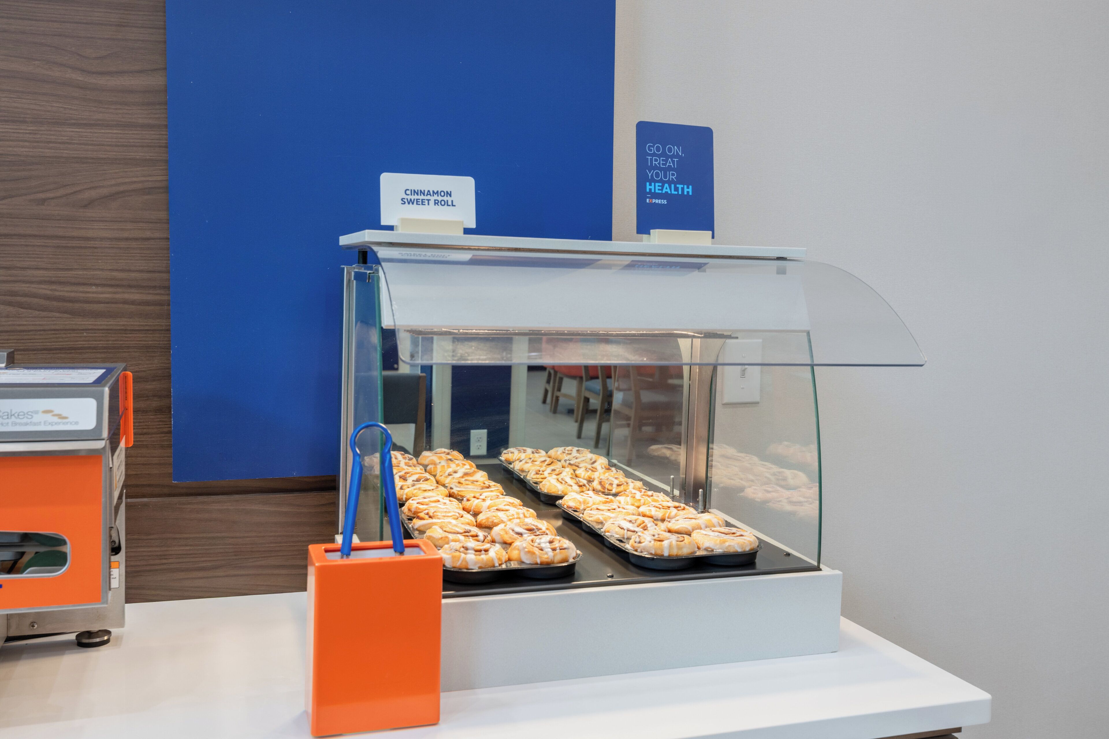 Een bakkerijvitrine met kaneelbroodjes en een bord waarop staat 'Ga gerust jezelf verwennen'.