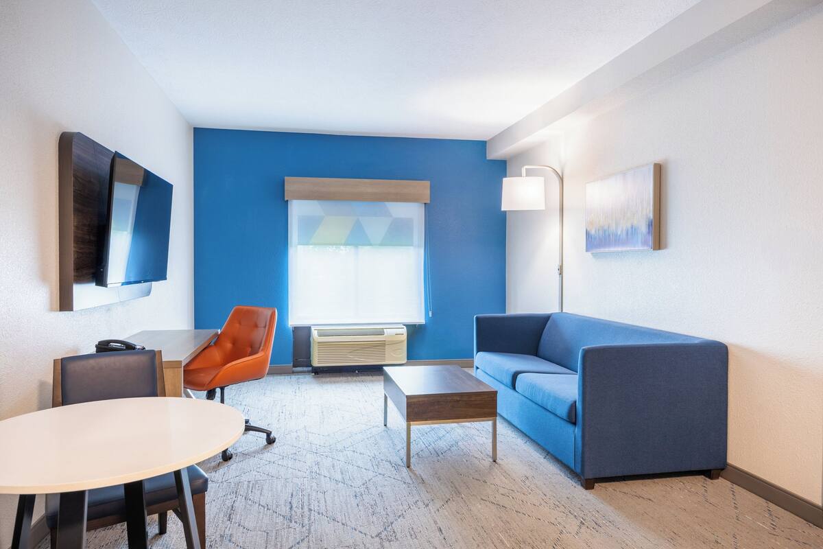 Camera d'albergo moderna con parete azzurra, un divano, un tavolo, una sedia, una scrivania, una lampada e un'ampia finestra.