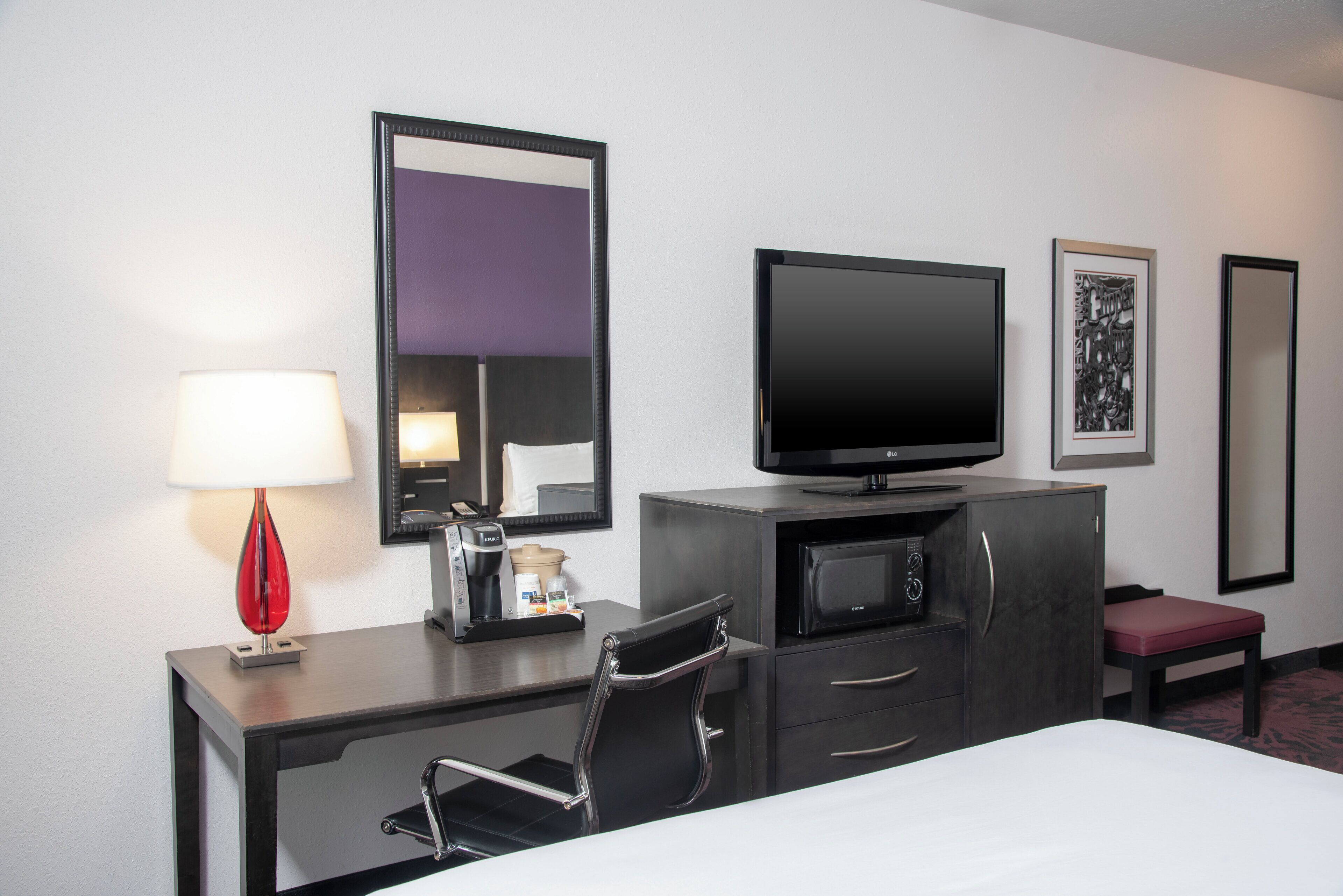 Ein Hotelzimmer mit Bett, Schreibtisch mit Lampe, Fernseher, Kaffeemaschine, Mikrowelle und Stuhl.