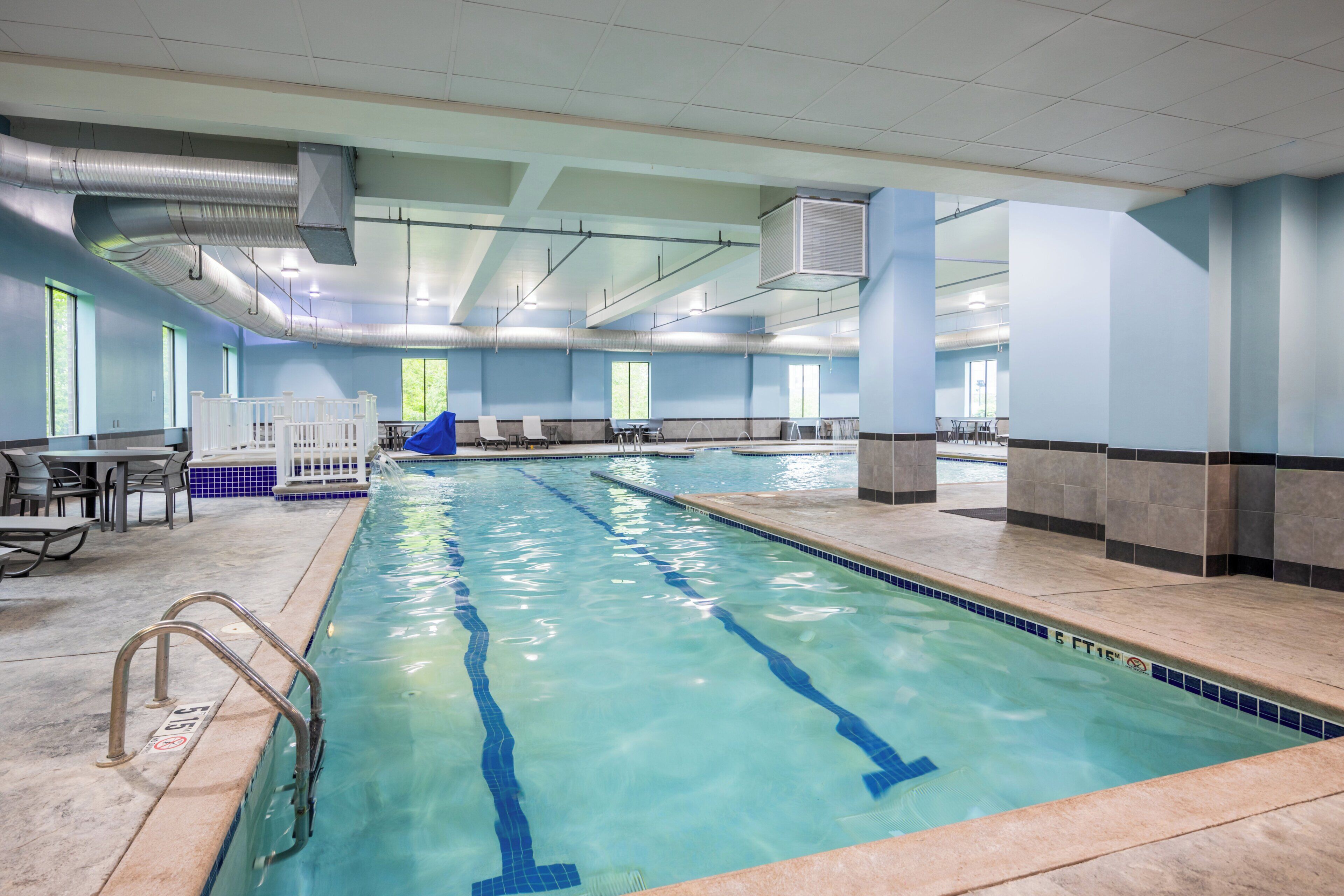 An indoor swimming pool with clear lane markings, a lifeguard chair, and a seating area with tables and chairs.