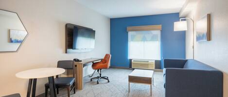A modern hotel room with a blue accent wall, a flat-screen TV, a sofa, a desk, and a small round table.