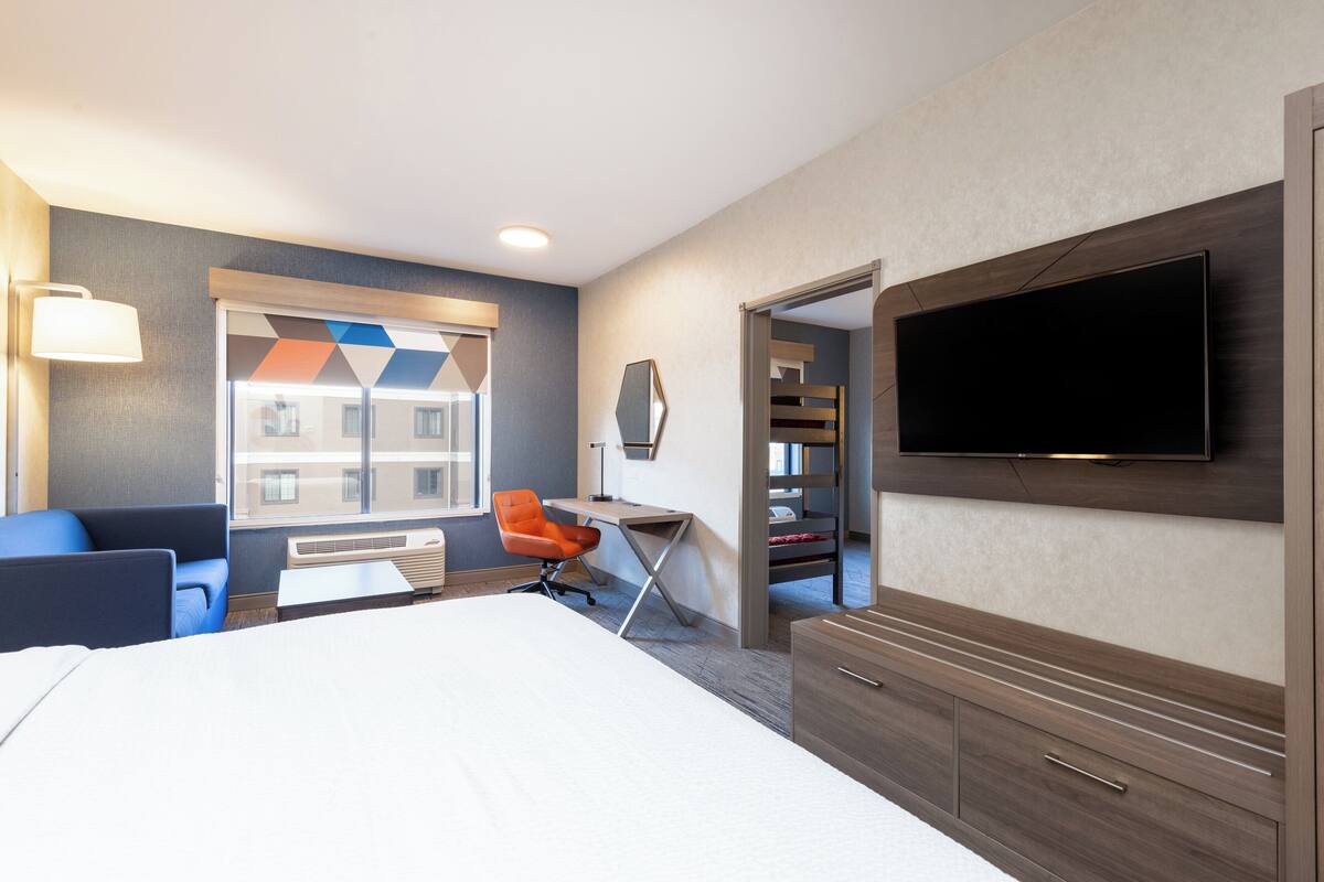 A hotel room with a bed, a desk, an orange chair, a TV, and a view of the outside.