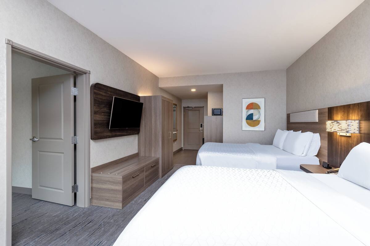 A hotel room with two beds, a TV mounted on the wall, a bedside table, and a wall-mounted lamp.