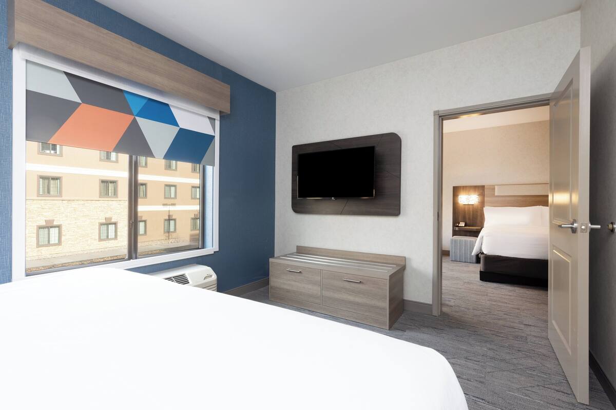 A hotel room with a bed, a TV mounted on the wall, a dresser, and a window with a geometric view.