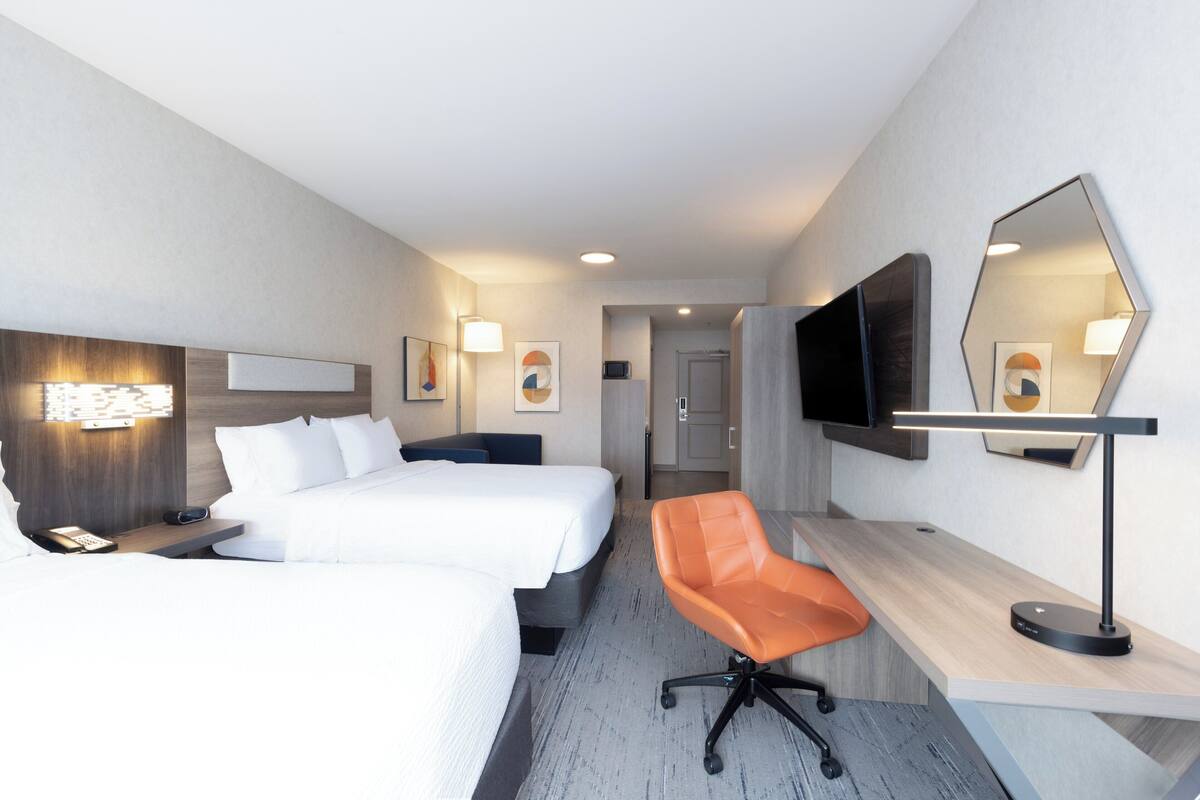A hotel room with two beds, a desk, an orange chair, a flat-screen TV, and a large mirror.