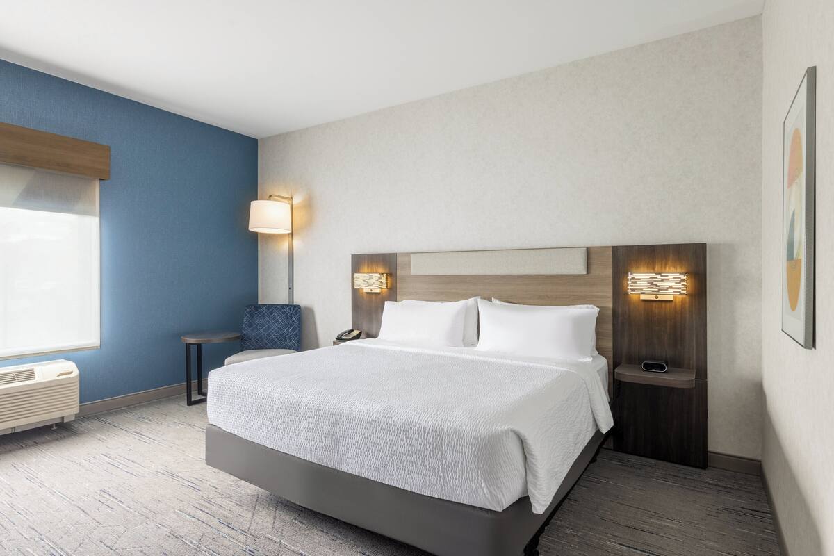 A hotel room with a large bed, a nightstand with a lamp, a chair, and a wall-mounted heater.