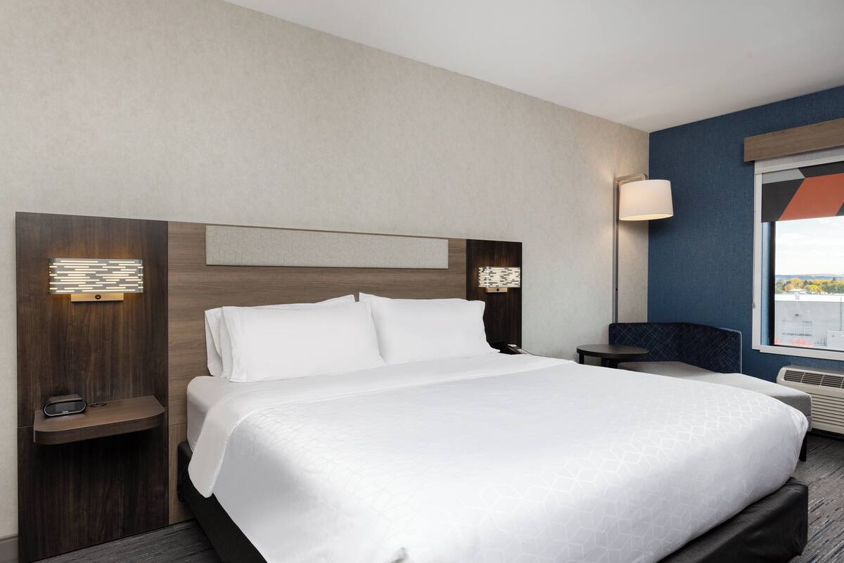 A modern hotel room with a large bed, a bedside table with a lamp, and a view of the city through a large window.
