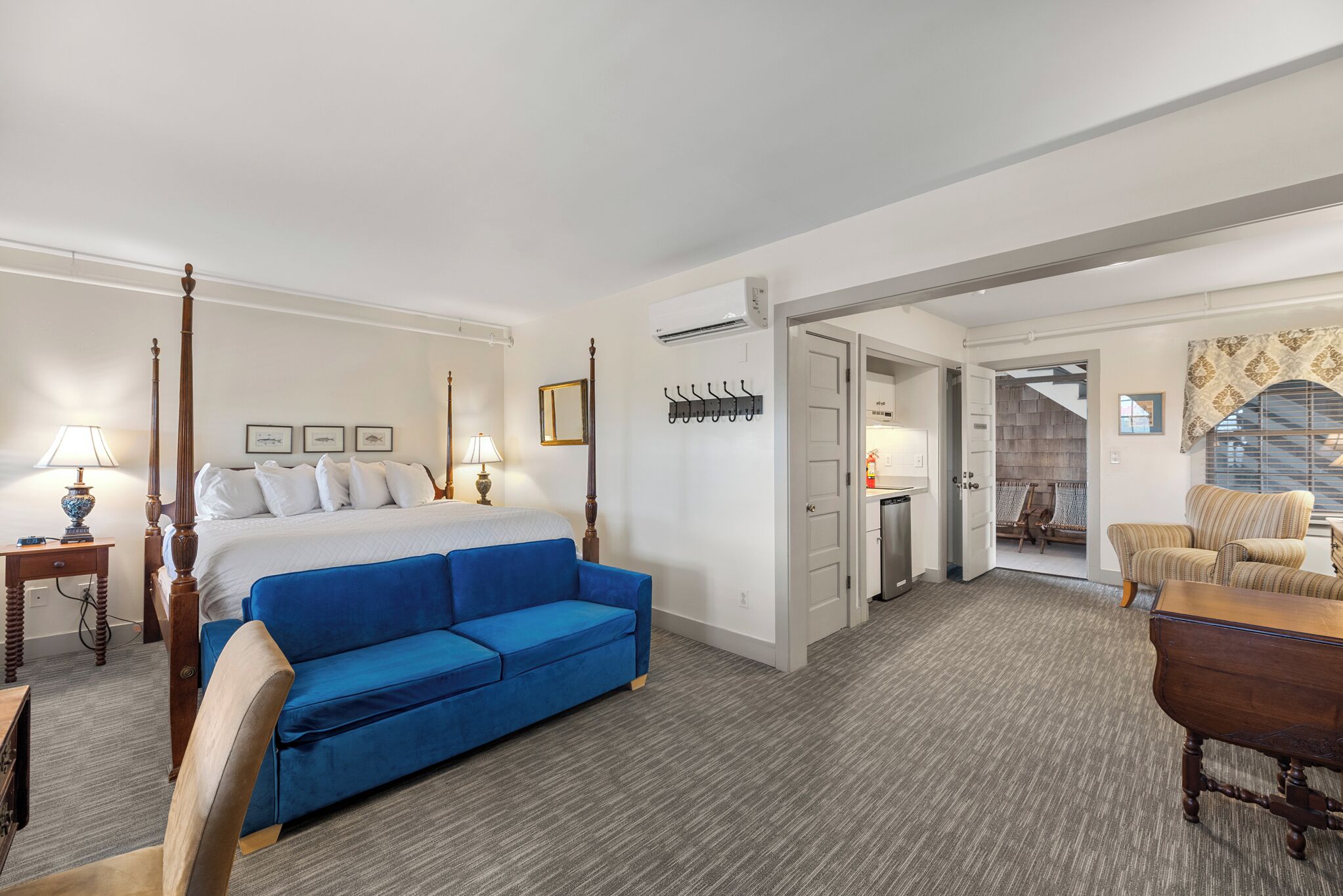 A hotel room with a large bed, a blue sofa, a wooden chair, a desk, and a small table. There is a coat rack with hangers, a lamp, and a picture frame on the wall.