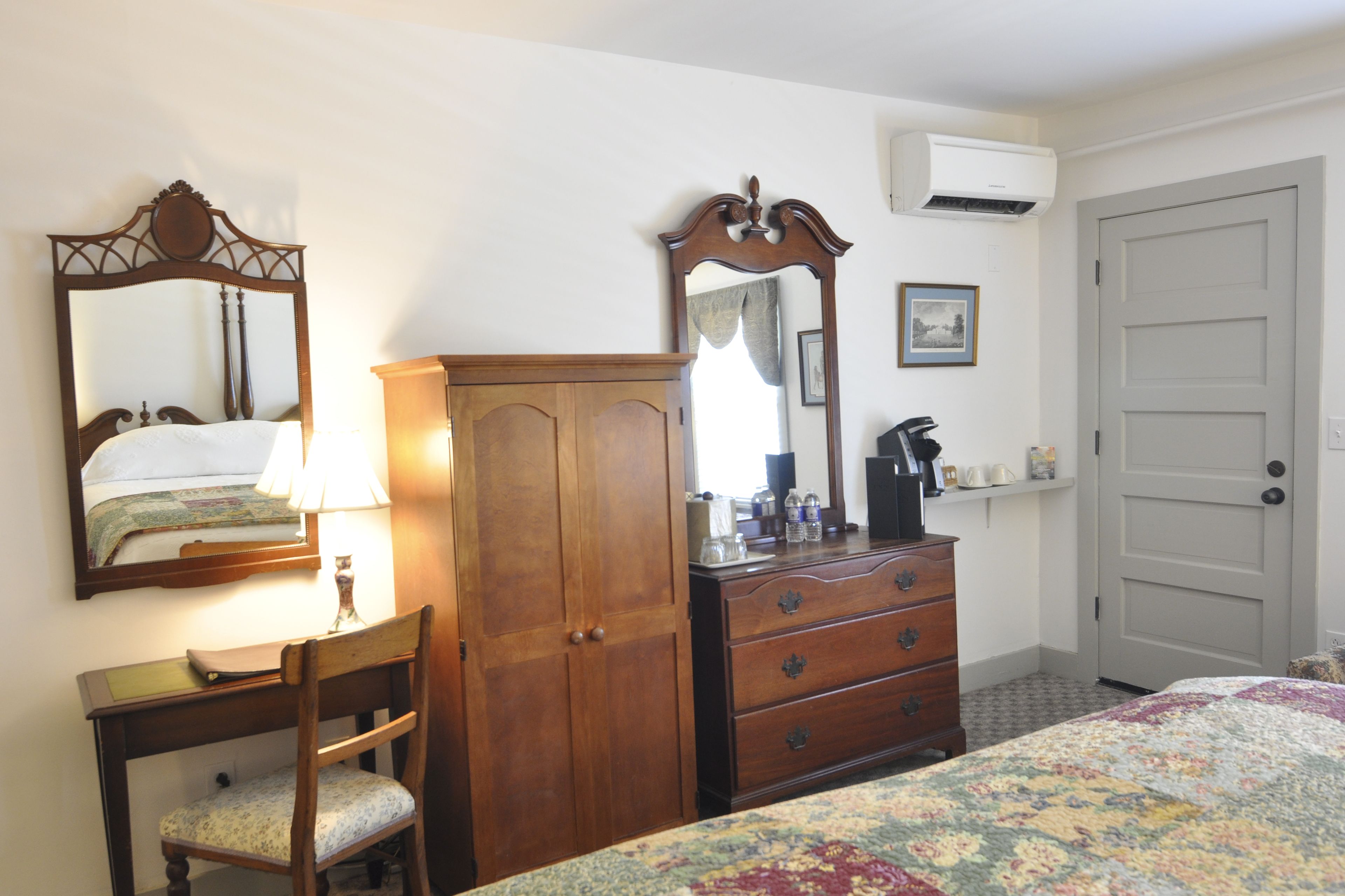 A bedroom with a bed, a wooden dresser, a desk with a chair, a mirror, and a door.