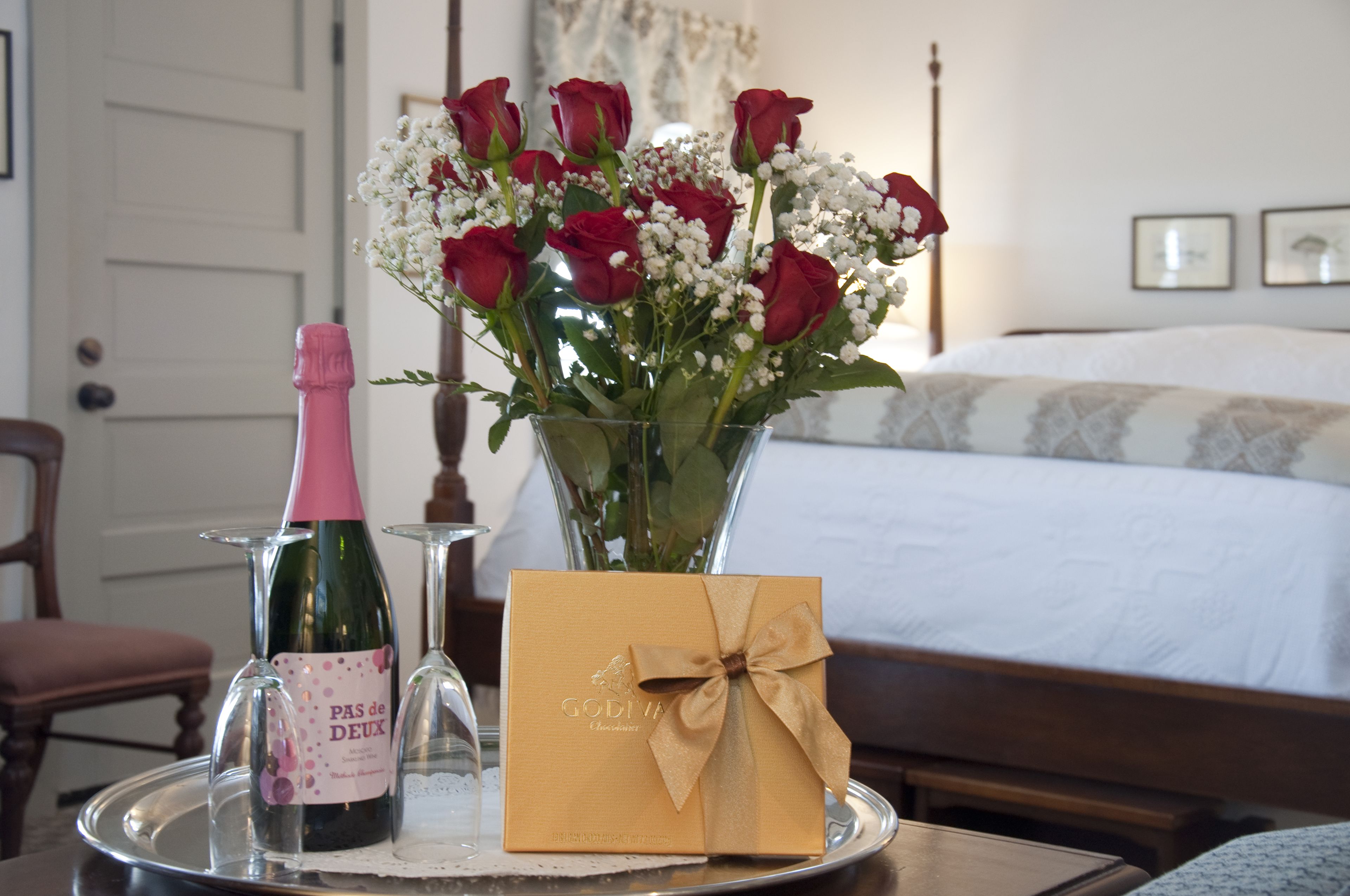 A bed, a bedside table, a vase with red roses, a bottle of champagne, and a gift box.