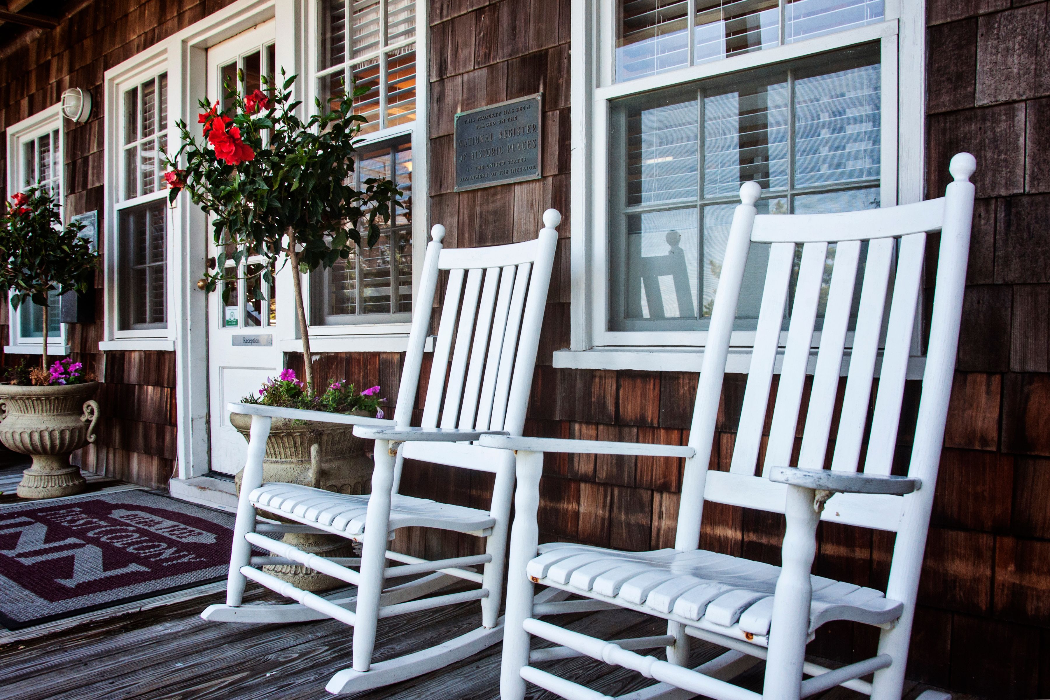 Two white rocking chairs on a wooden deck with a potted plant and flowers.