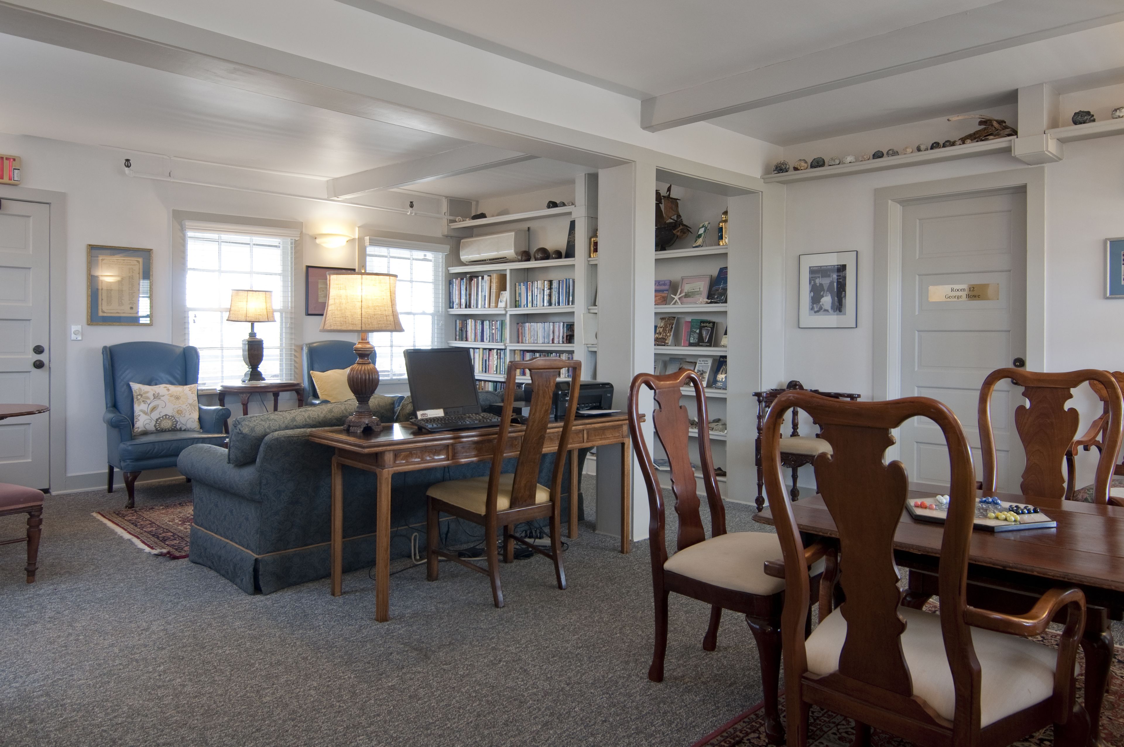 Un salon bien éclairé, avec un canapé bleu, des chaises en bois, une table à manger et une bibliothèque remplie de livres.