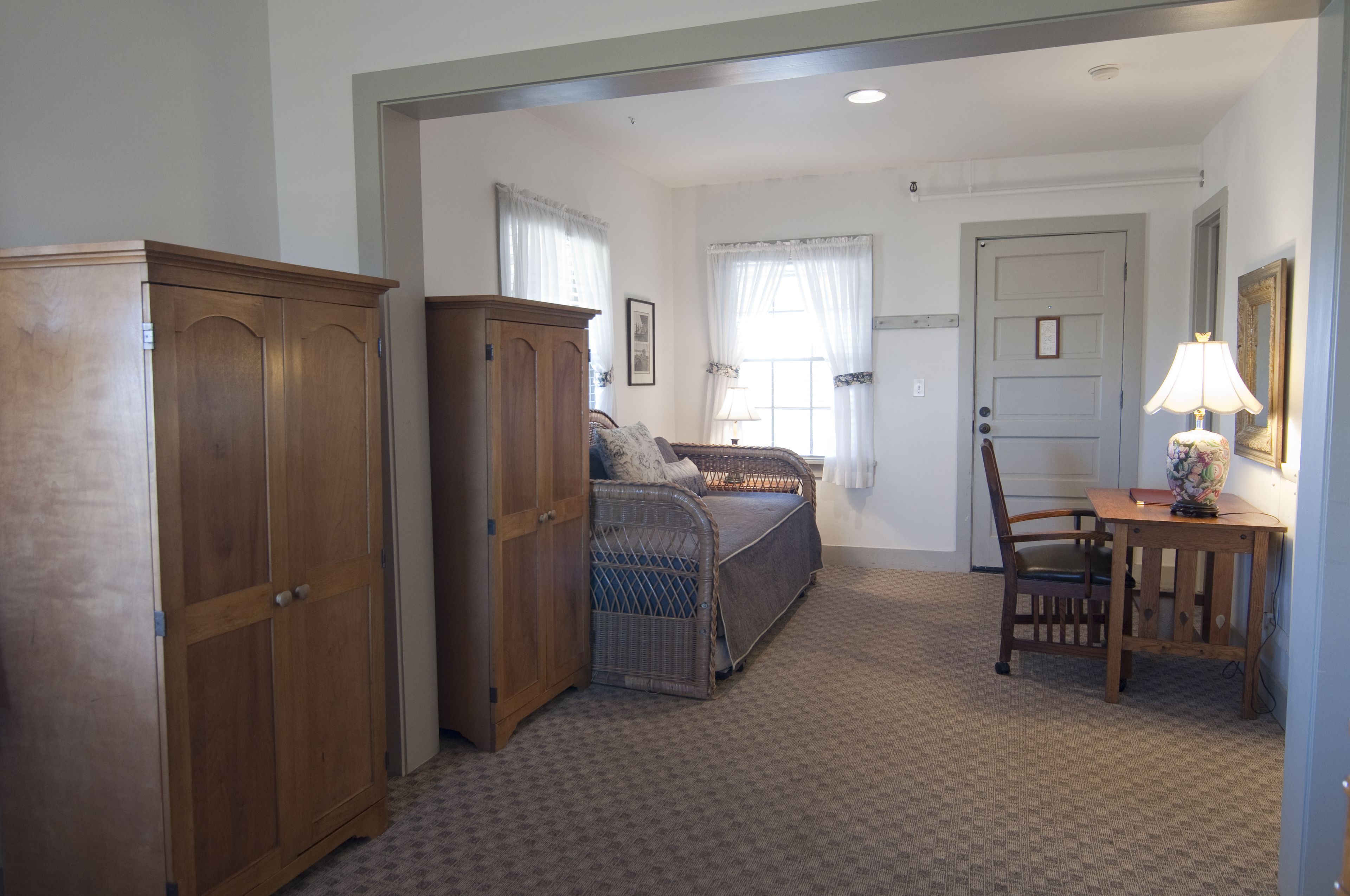 A bedroom with a bed, a wooden wardrobe, a wicker chair, a small table with a lamp, and a door.